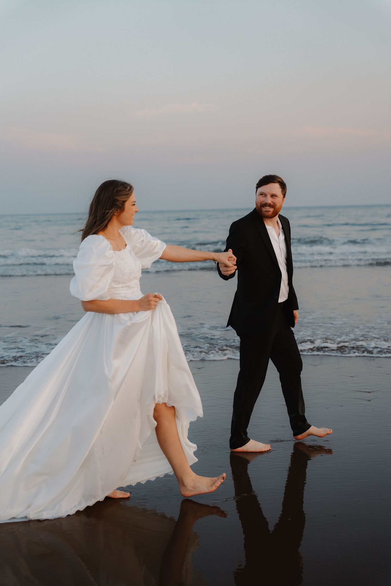 Barefoot bride and groom holding hands, dancing on a reflective beach at sunset in wedding attire.