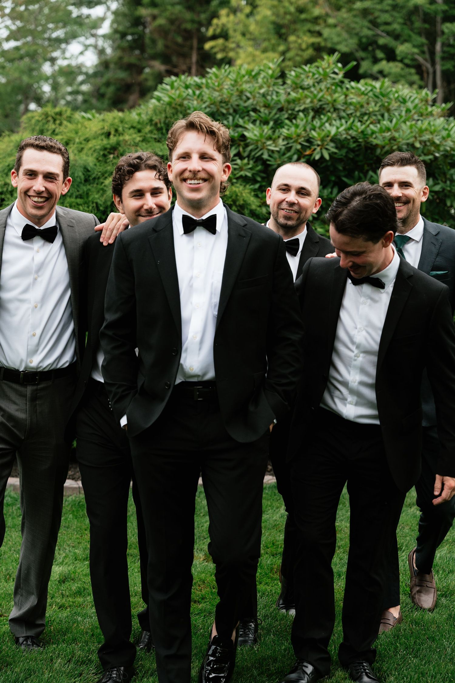 Smiling groom in black tuxedo posing with four groomsmen in matching suits on green lawn.