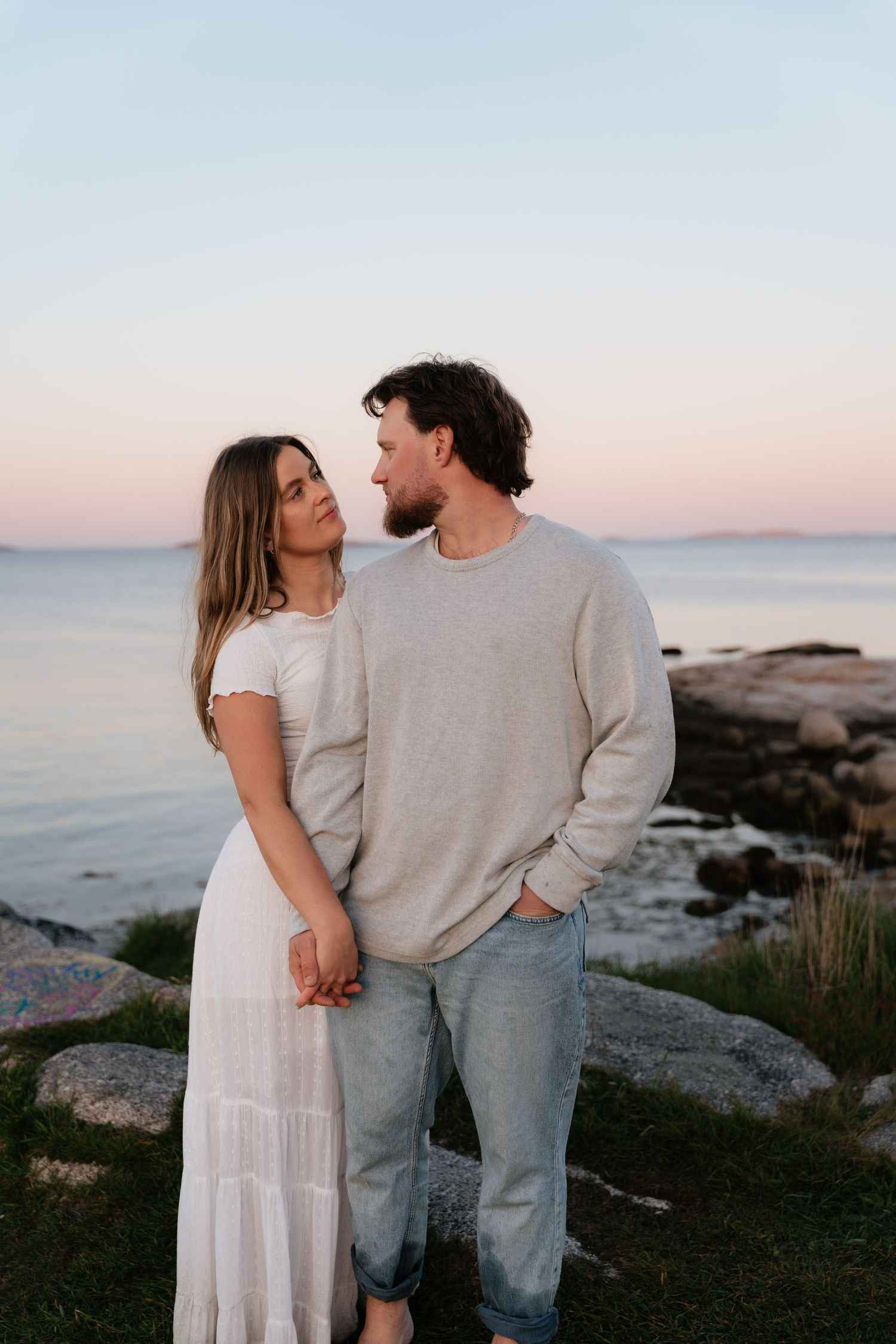 Couple holding hands and gazing at each other by the ocean at sunset, woman in white dress, man in beige sweater.