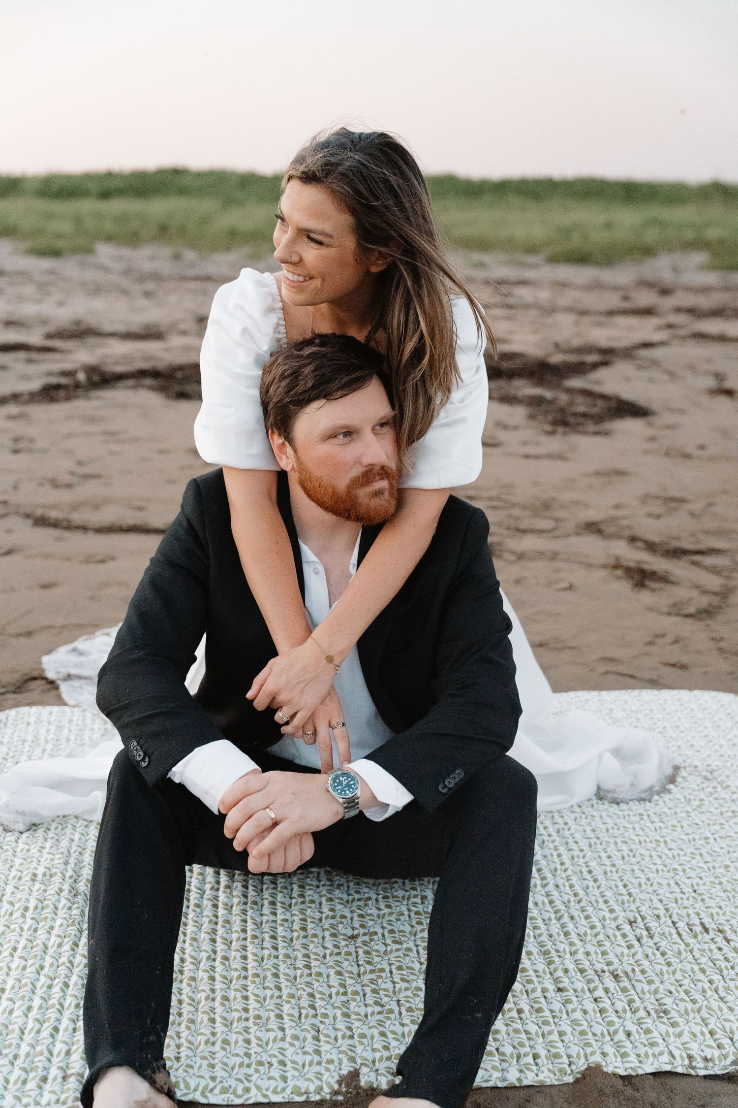 A couple sits on a blanket on a sandy beach, woman in white embracing man in black suit at dusk.