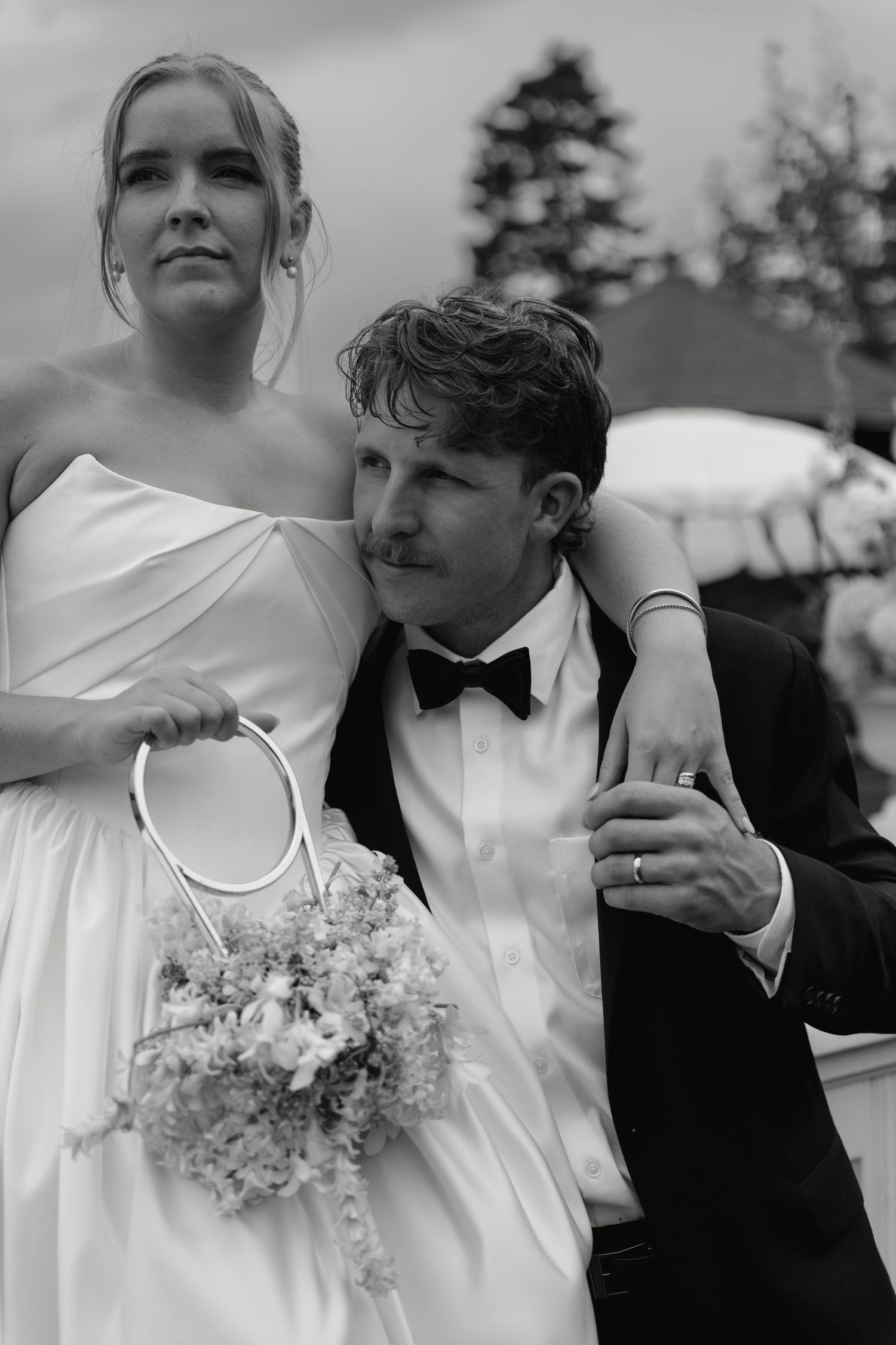 Black and white photo of bride in white gown and groom in tuxedo posing together at outdoor wedding.