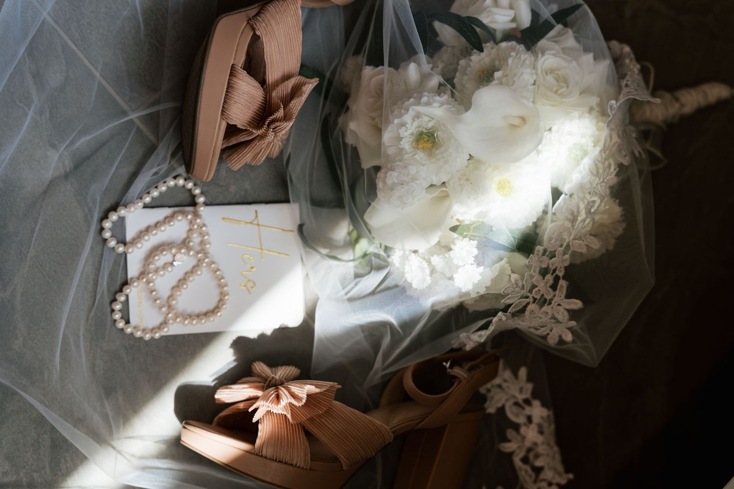 Elegant wedding flat lay with white peony bouquet, pearl necklace, blush heels, and sheer veil in soft natural light.