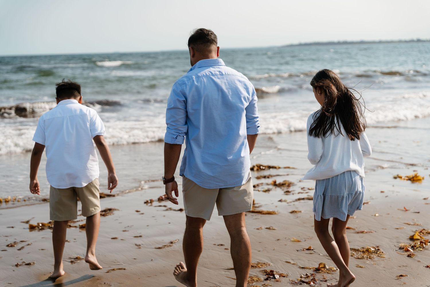 Family of three walking barefoot on a sandy beach toward ocean waves on a sunny day.