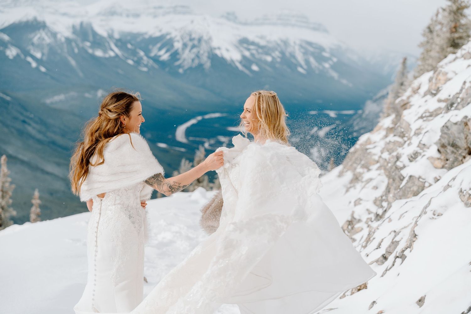 Two people in flowing white dresses dance on a snowy mountain peak with dramatic views.