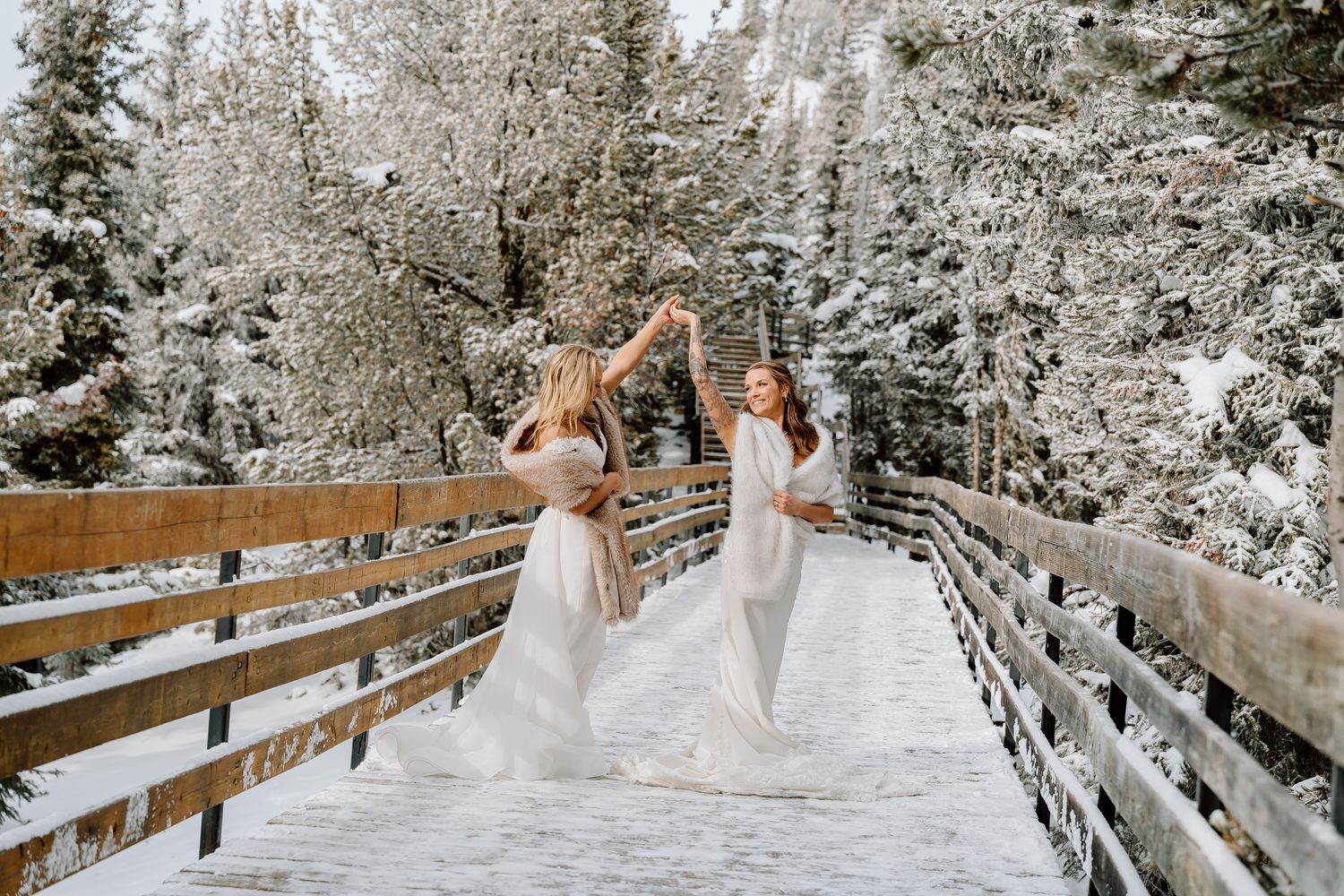 Two people in white dresses walk along a snow-covered wooden bridge surrounded by frost-covered evergreen trees.