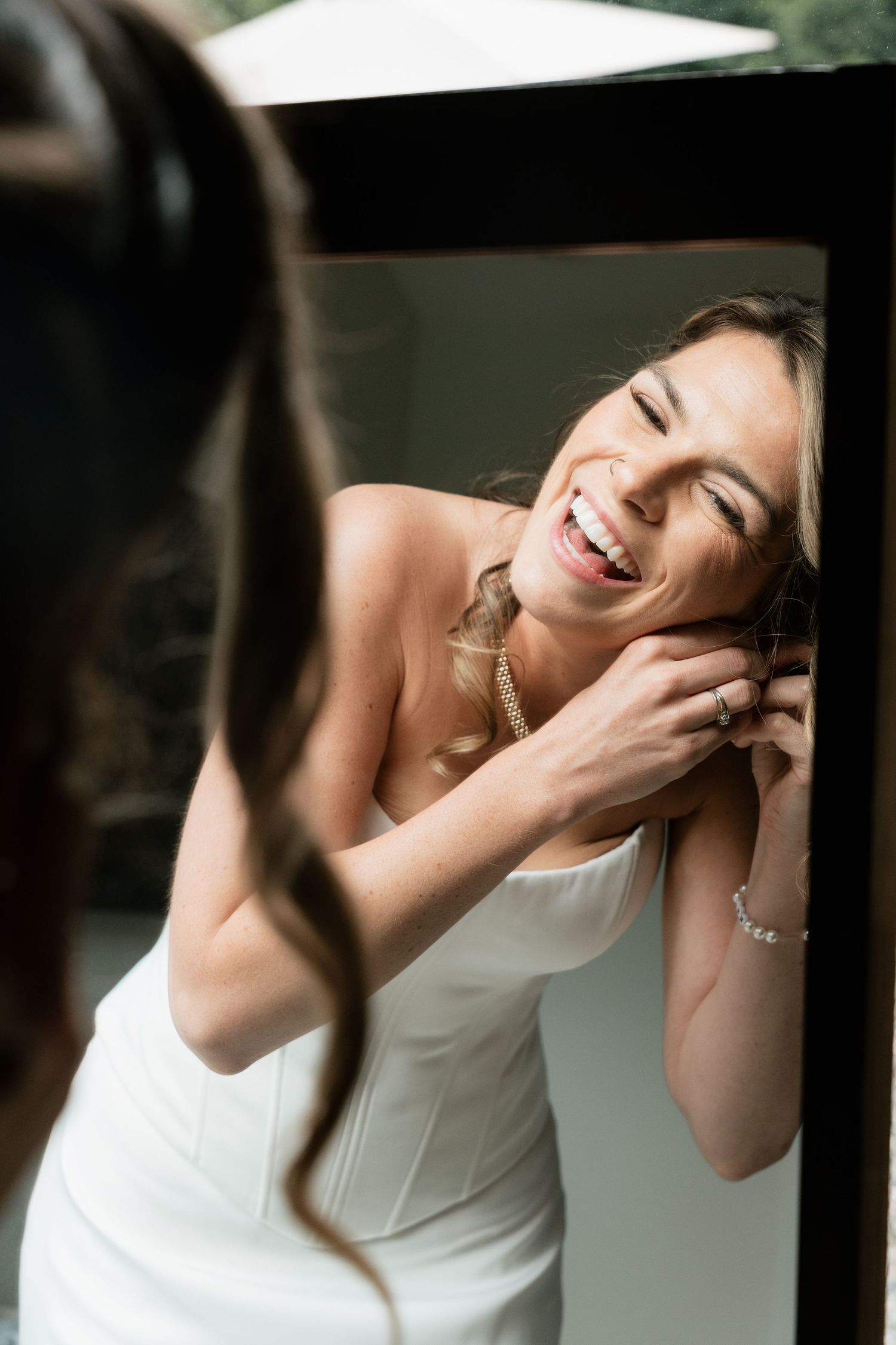 Smiling bride in white strapless gown putting on earrings while looking in mirror on her wedding day.