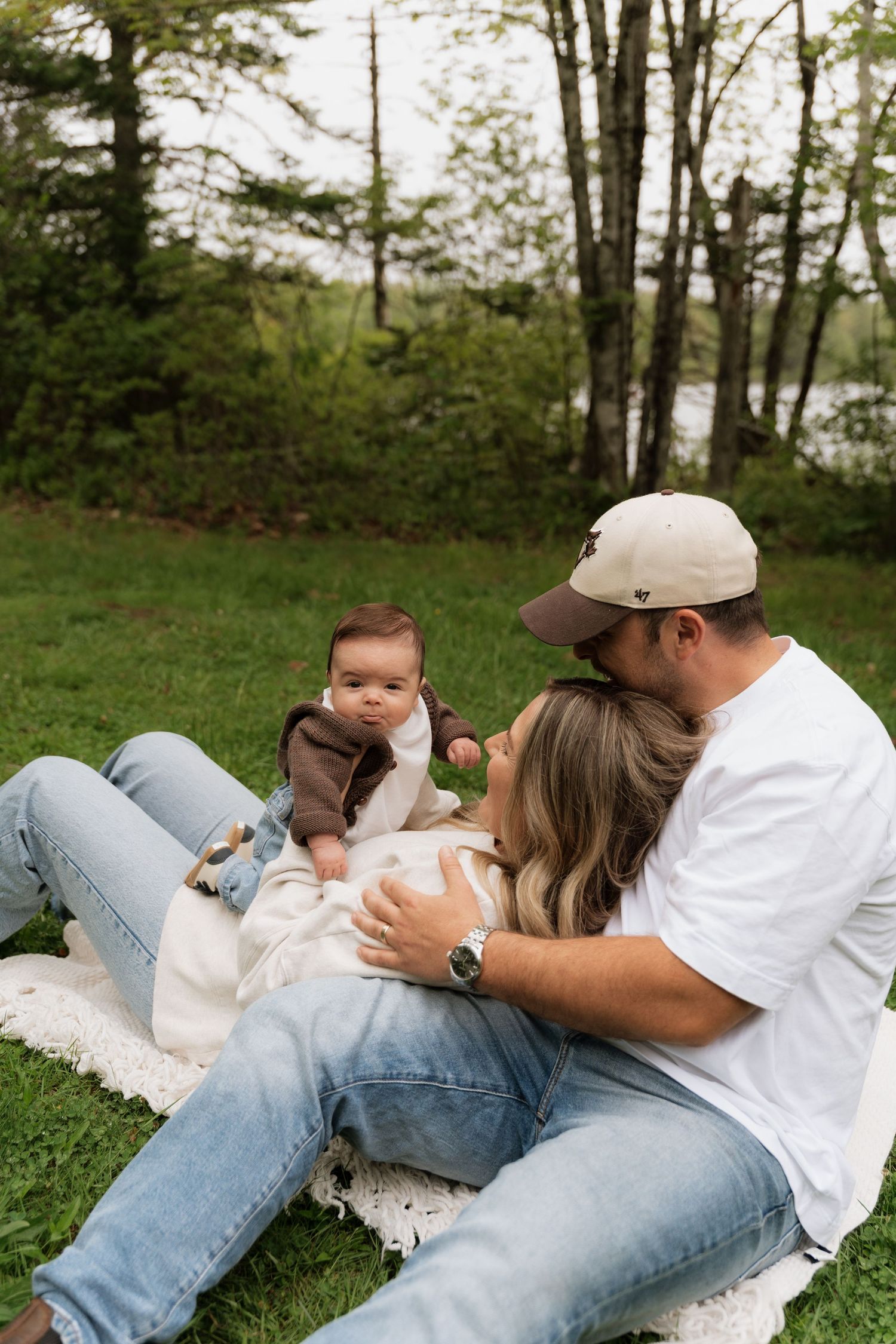 Family relaxing on a blanket in a green park, parents kissing while holding their baby outdoors.