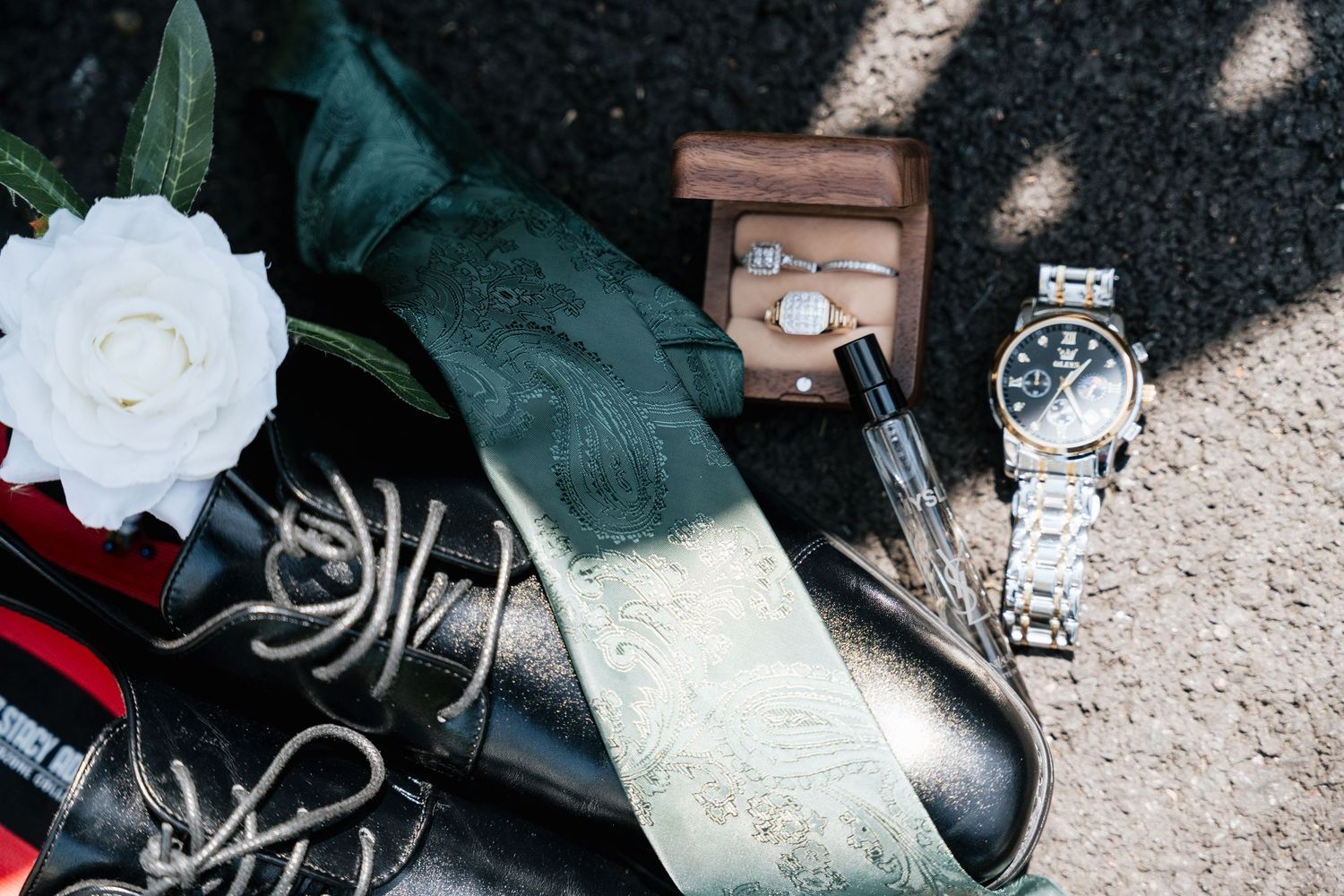 Groom's wedding accessories flat lay featuring a green tie, dress shoes, watch, ring box, and white rose.