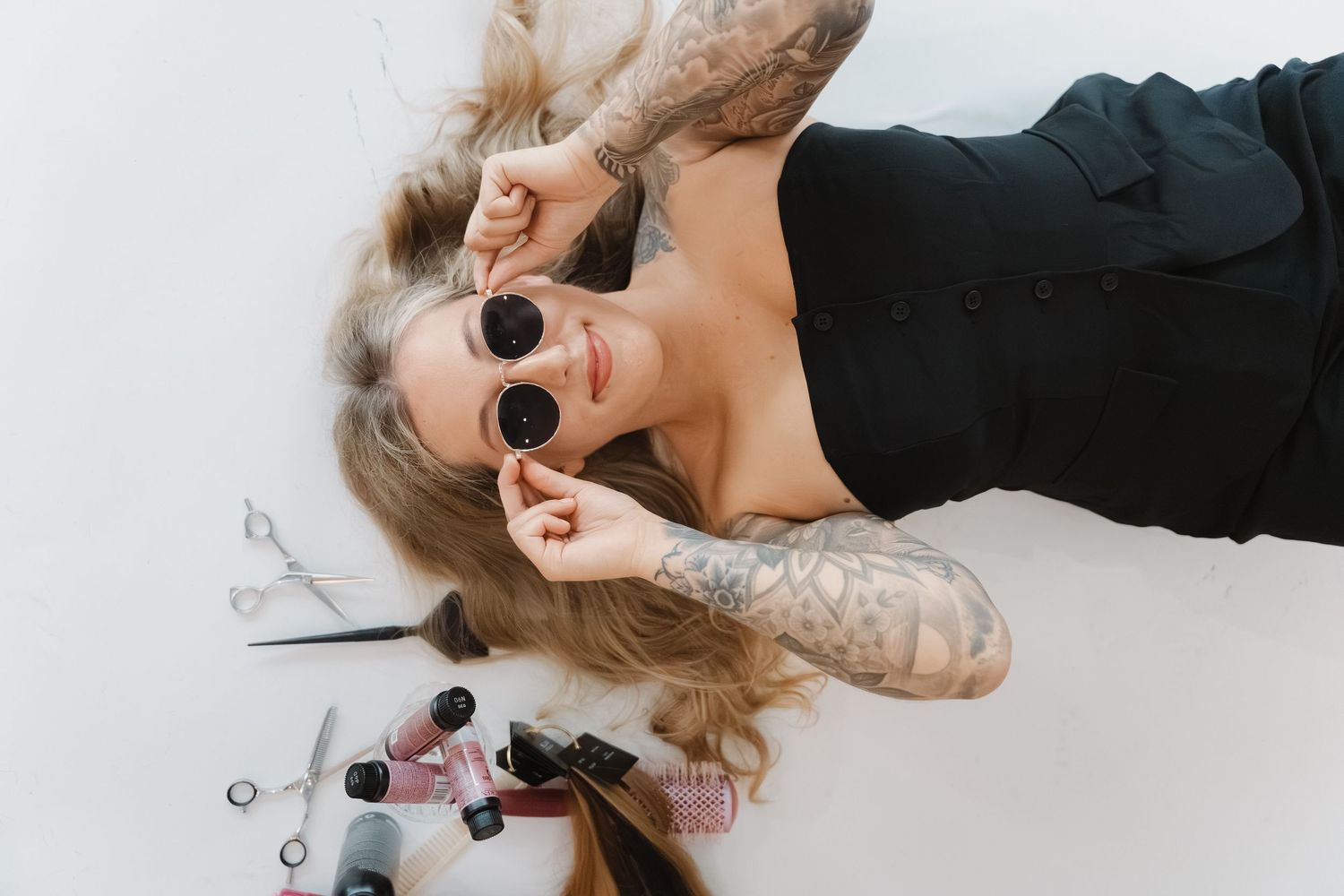 Tattooed woman lying on floor wearing black outfit and round sunglasses, surrounded by nail polish bottles and scissors.