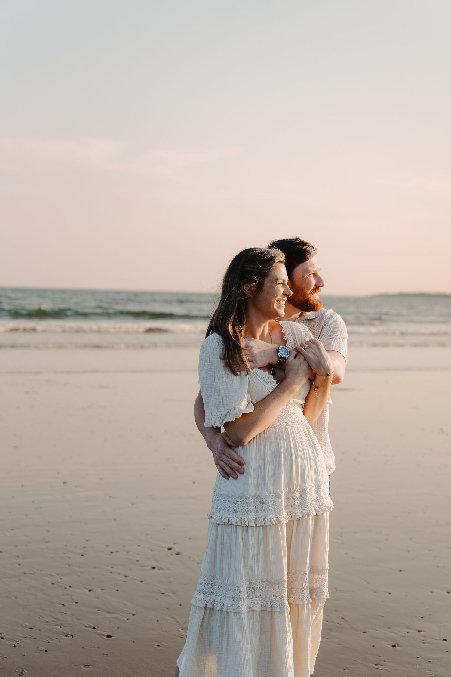 Couple embracing on a beach at sunset, woman in white boho dress smiling joyfully with ocean waves in background.