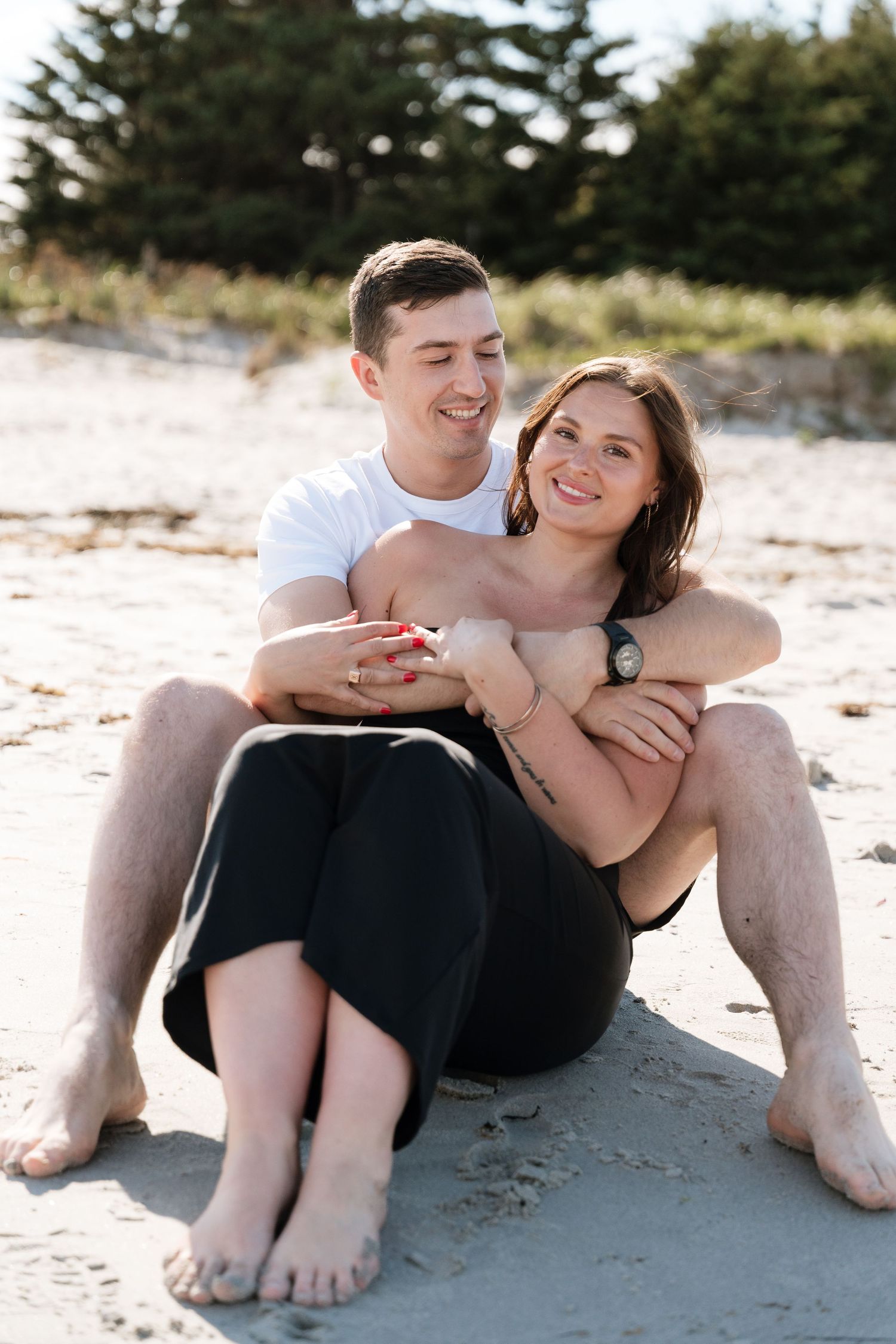A happy couple sits together on a sandy beach, the man embracing the woman from behind, both smiling warmly.