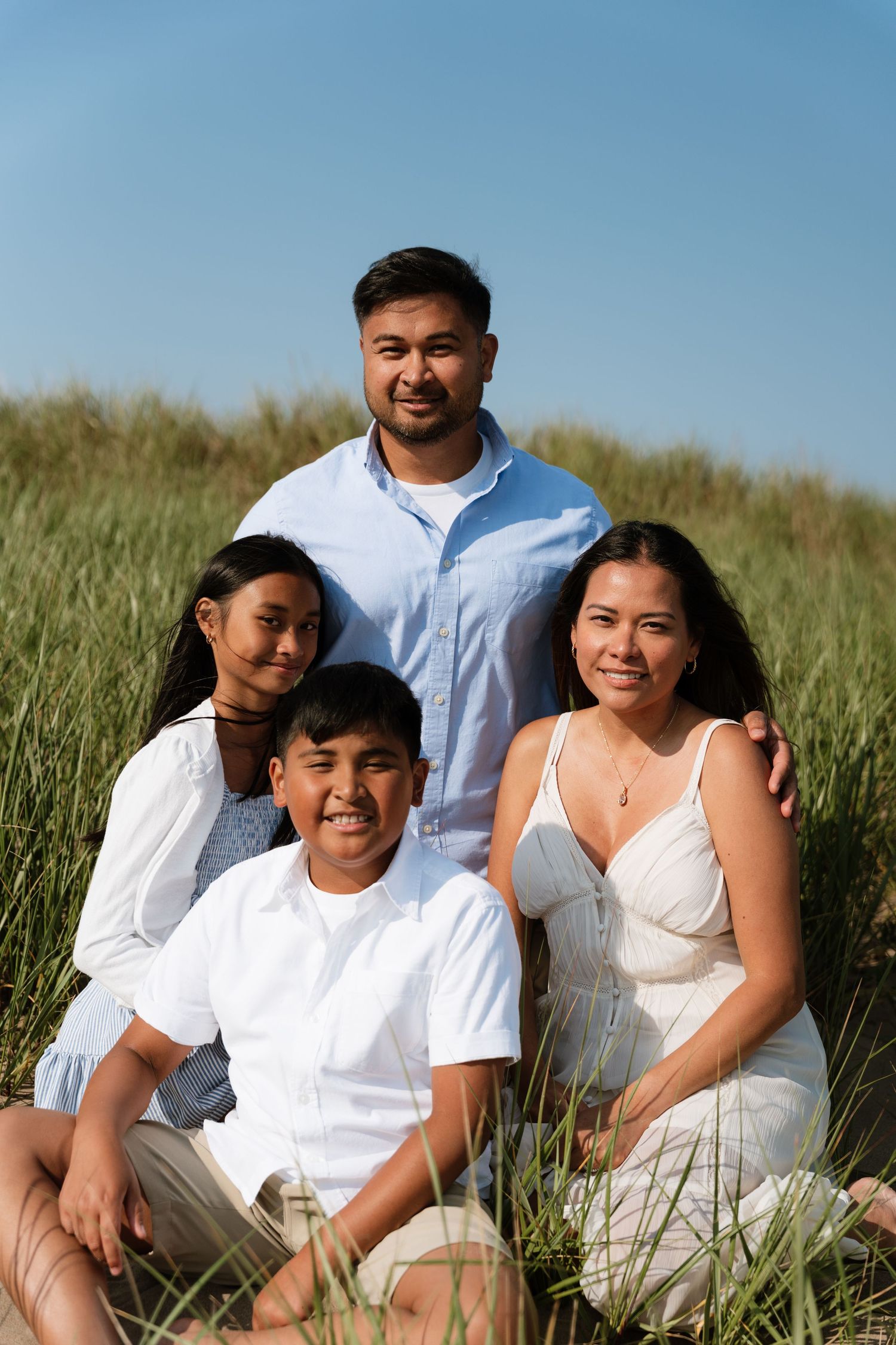 A happy family of four poses in green dunes under a blue sky, wearing white and light blue outfits.