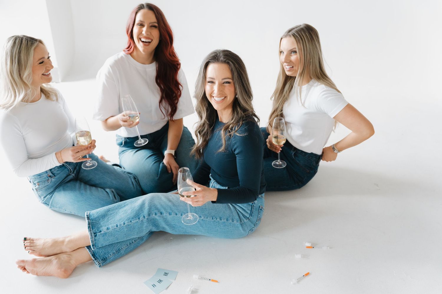 Four women in white tops and jeans sitting on floor holding wine glasses, smiling and celebrating together.
