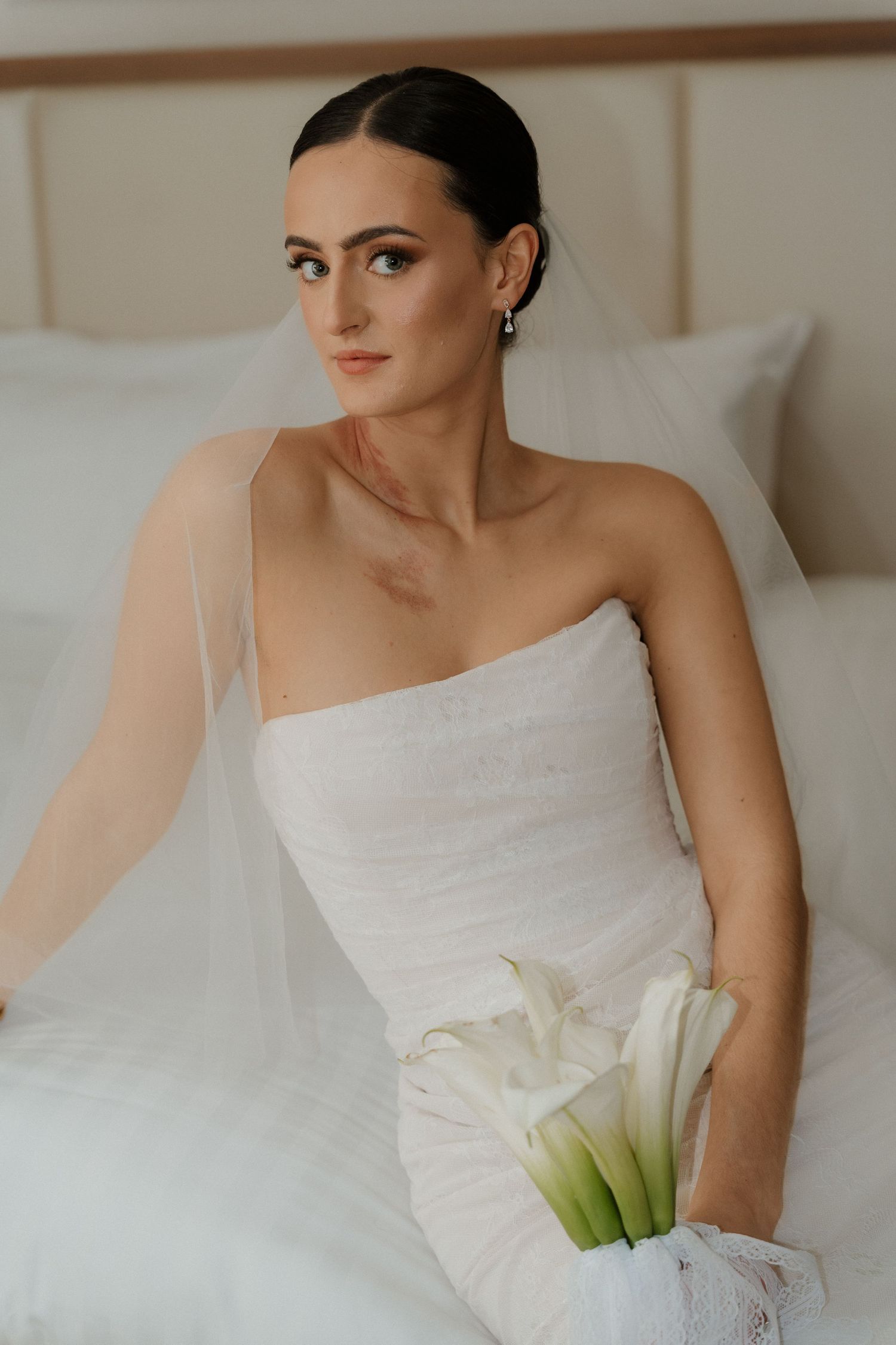 Elegant bride in white strapless wedding gown holds calla lily bouquet while sitting on bed with soft lighting.