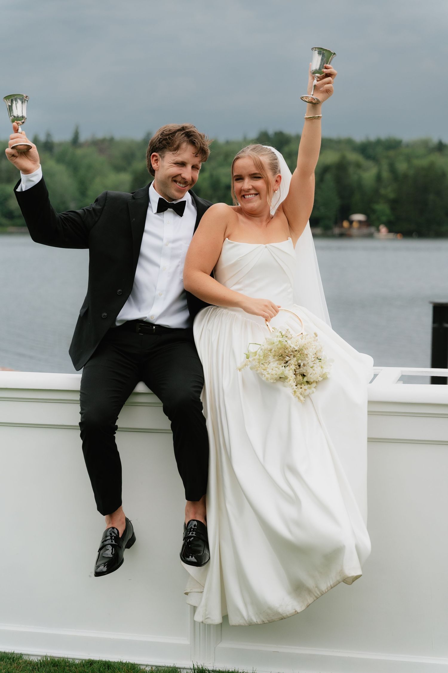 Joyful bride and groom raising champagne glasses outdoors by a lake, bride in white gown holding bouquet.