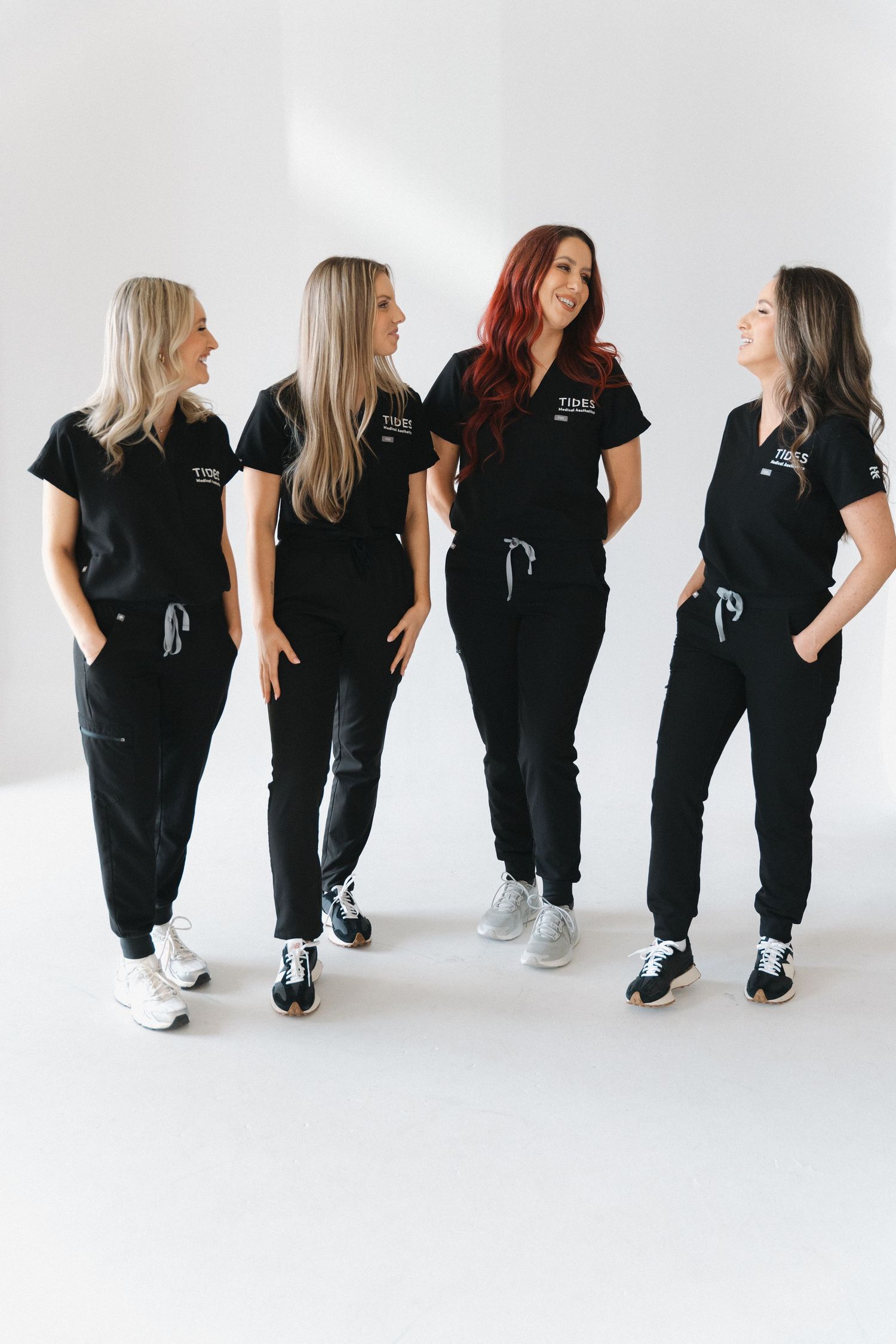 Four women in matching black scrubs smiling and posing together against a white background.