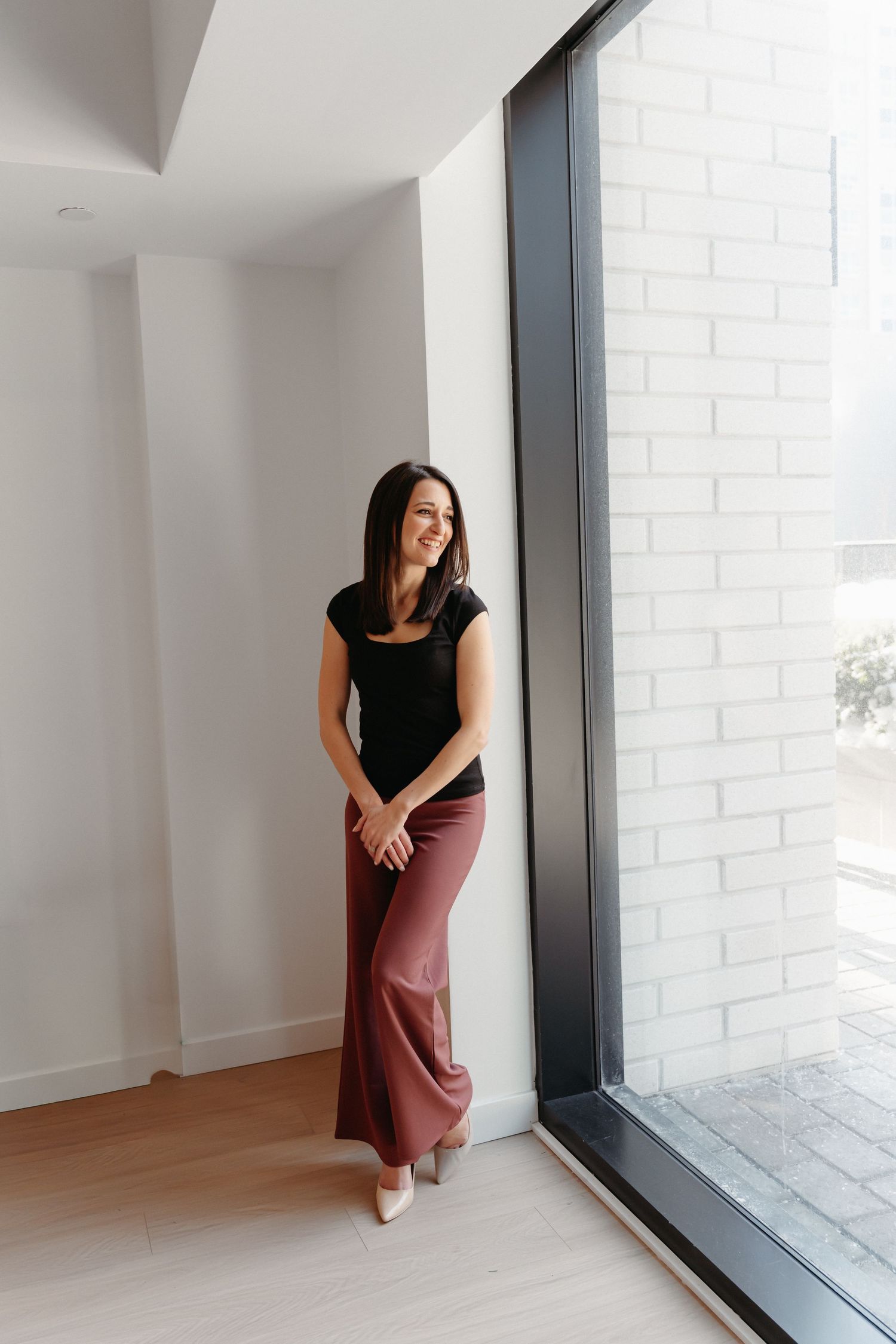 A smiling woman in a black top and mauve wide-leg pants leans against a white wall near a large modern window.