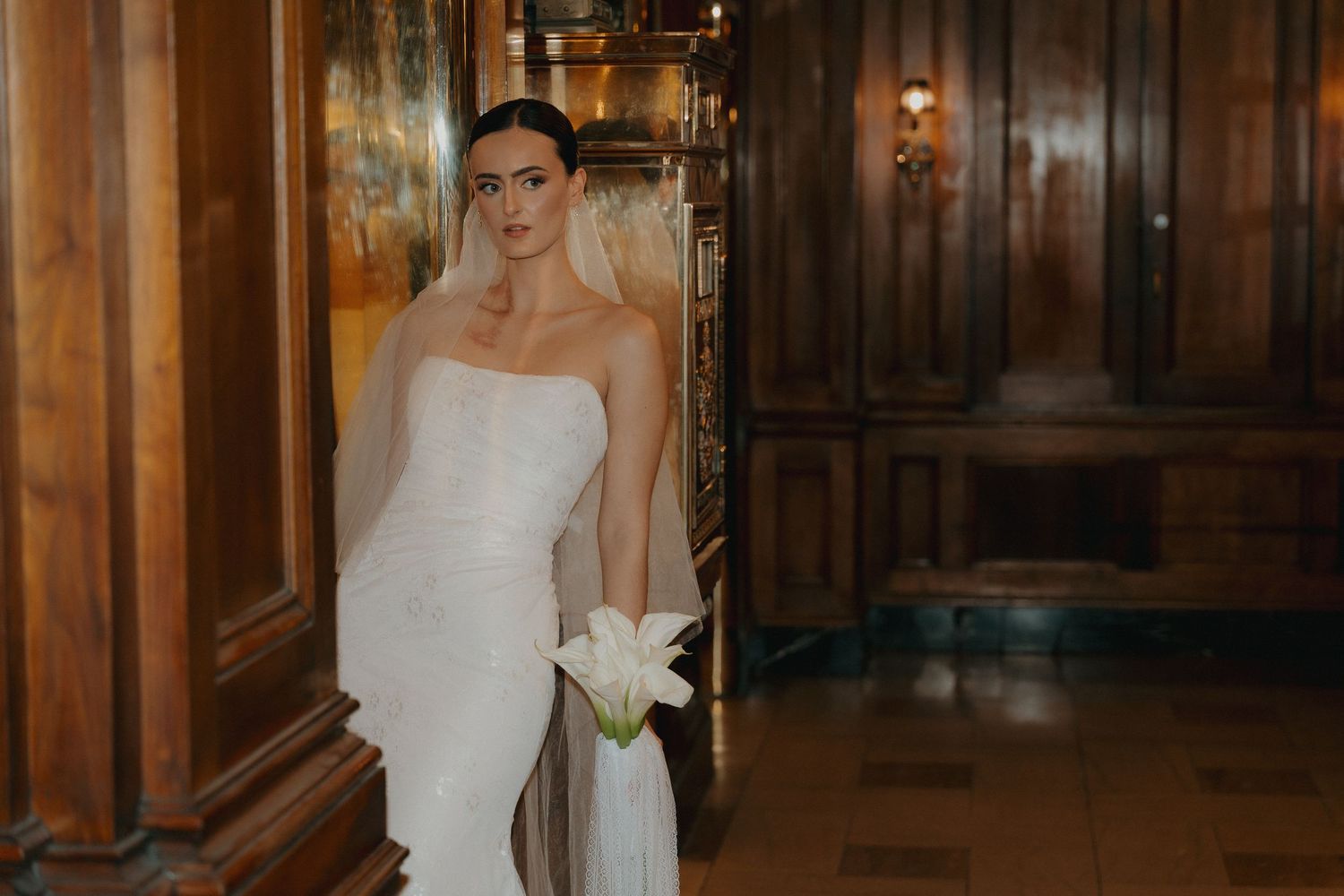 Elegant bride in strapless white gown holds lily bouquet while standing in wood-paneled hallway.