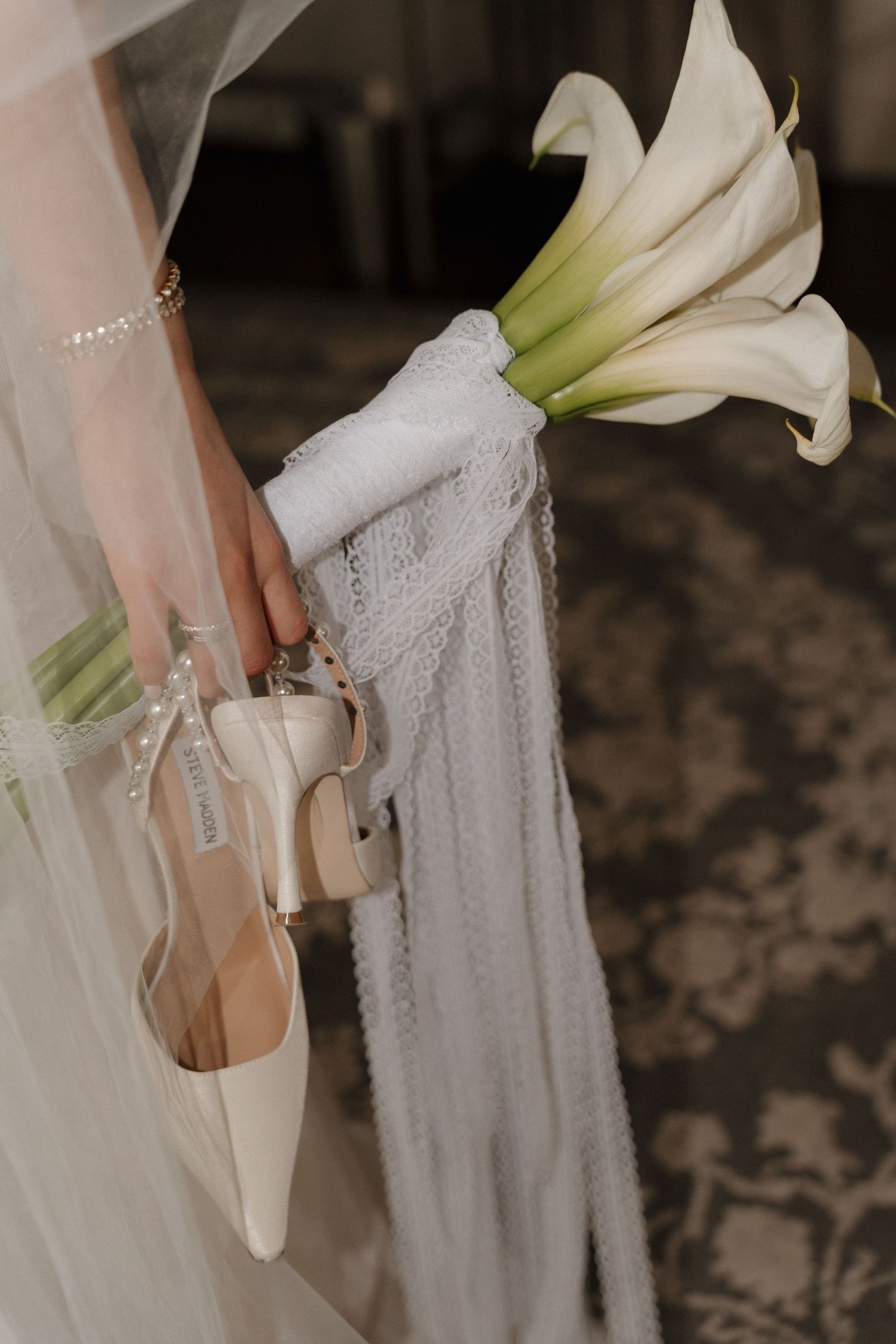 A bride holds white calla lilies against flowing lace fabric in an elegant wedding detail shot.