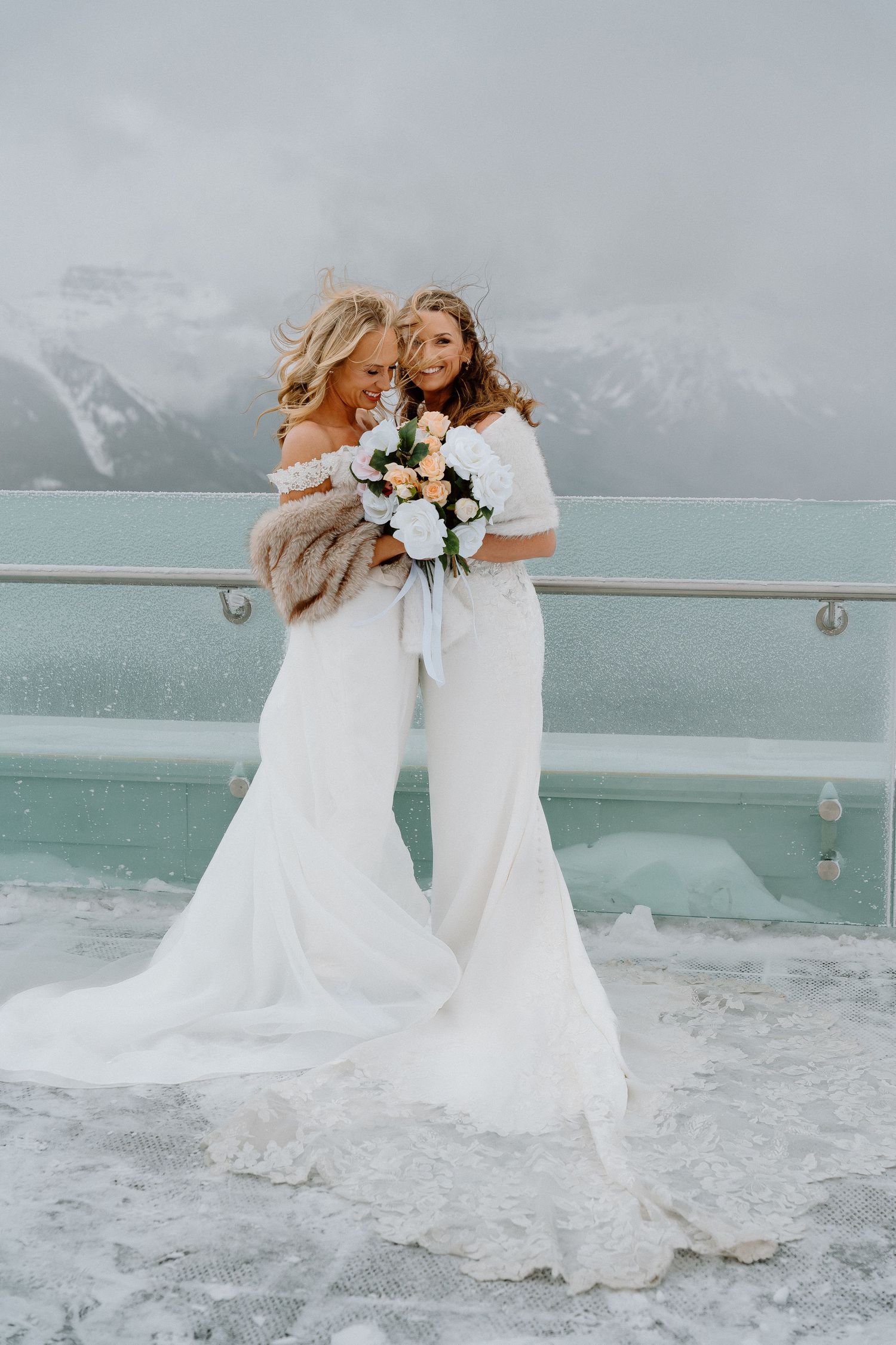 Two brides in white dresses pose together against snowy mountain landscape and turquoise lake.