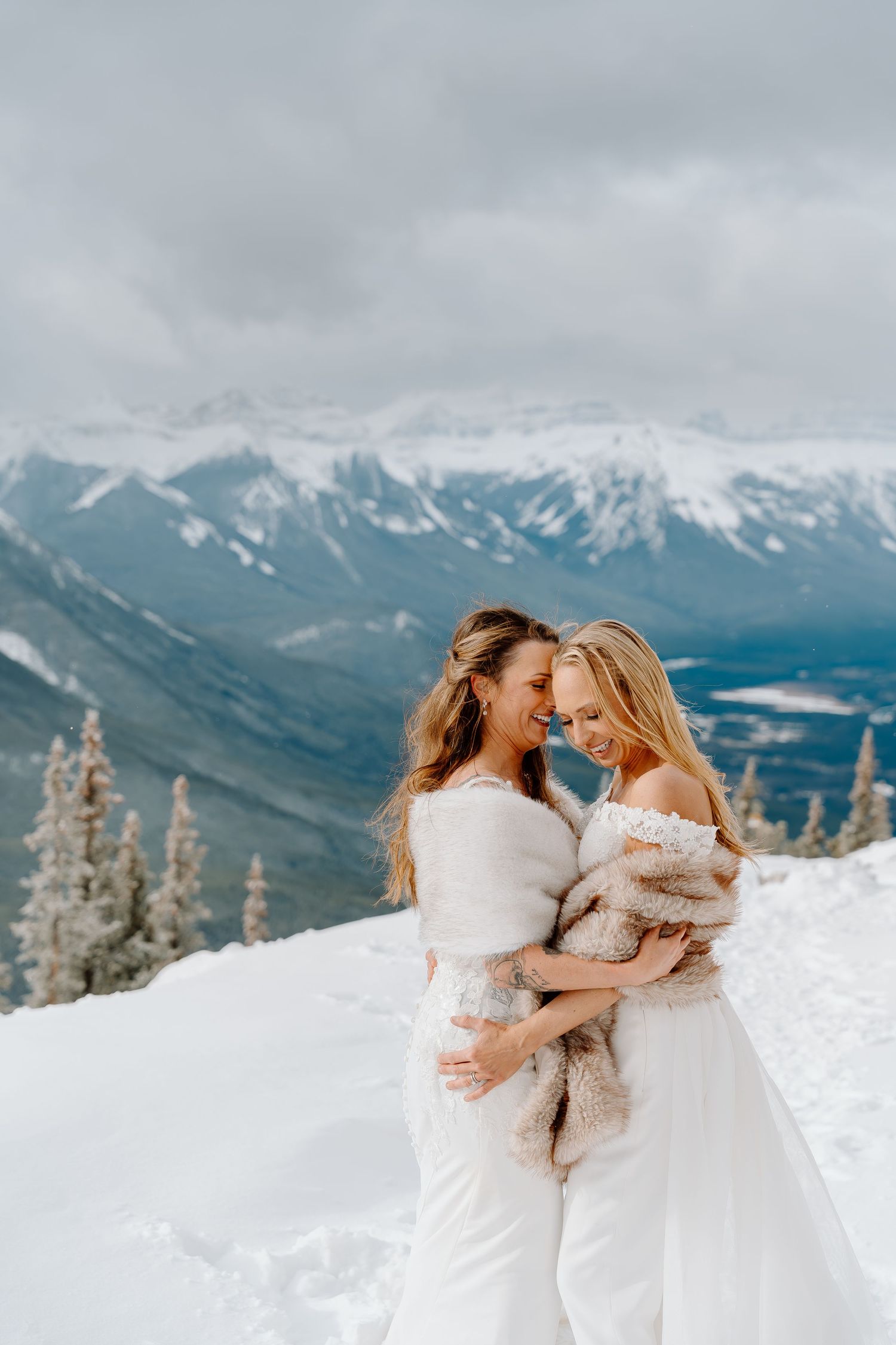 Two people in white winter outfits embrace on a snowy mountain peak with dramatic snow-capped mountains in background.