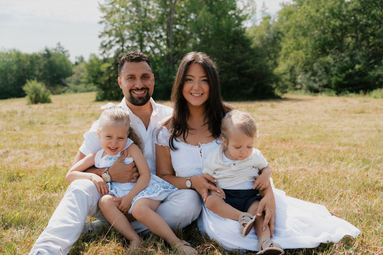 Happy family of four sitting on grass in a sunny park, parents smiling with two young children on their laps.
