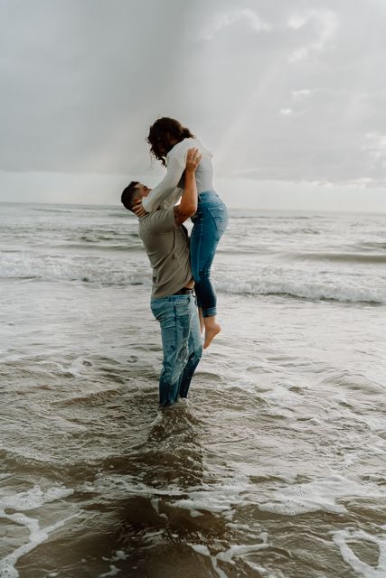 Playful couple splashes in ocean waves during moody beach photoshoot.
