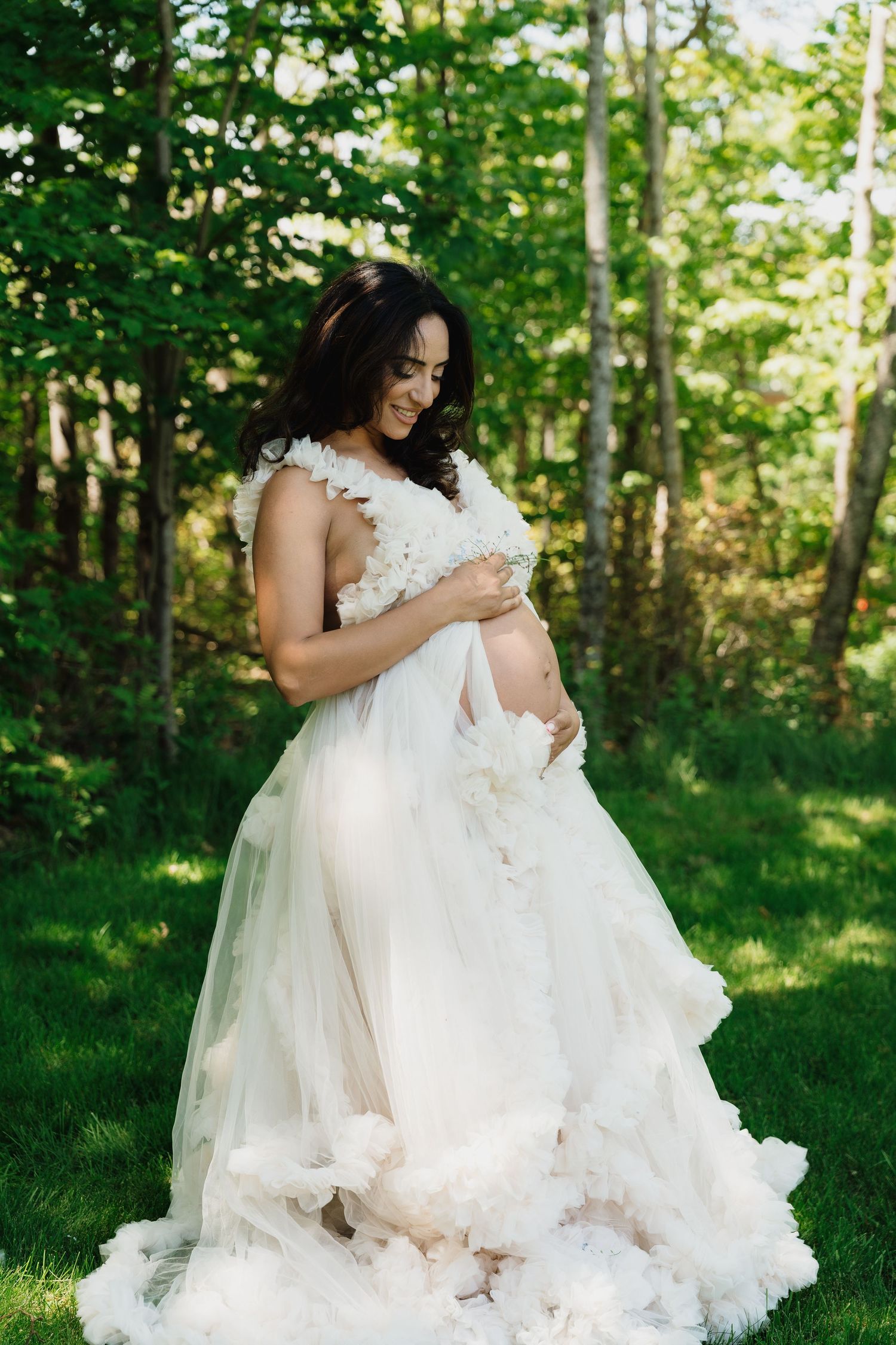 Pregnant woman in white tulle gown holding baby bump outdoors in lush green forest setting.