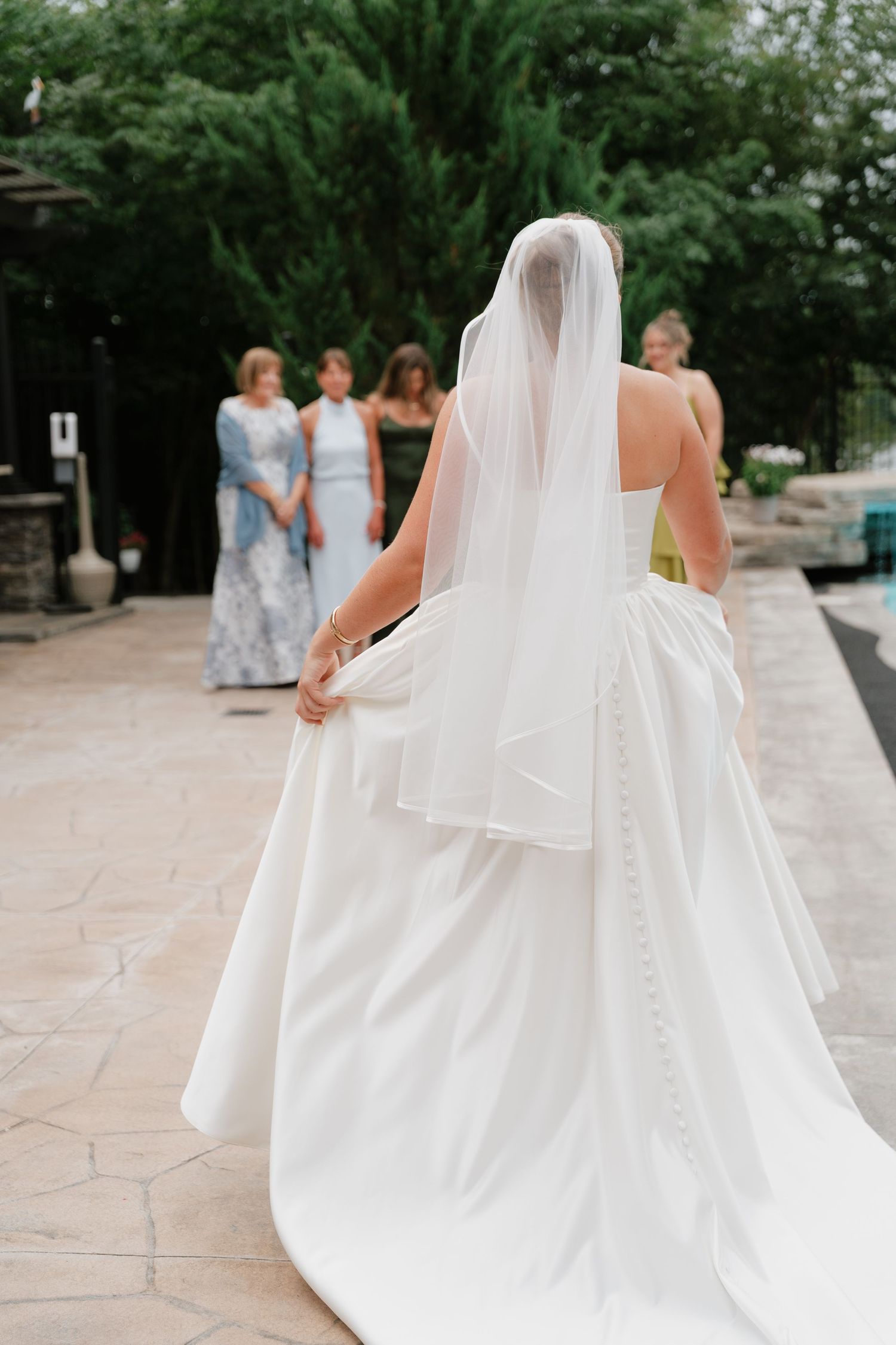 Bride in strapless white gown with veil and button-back train walking toward bridesmaids on patio.