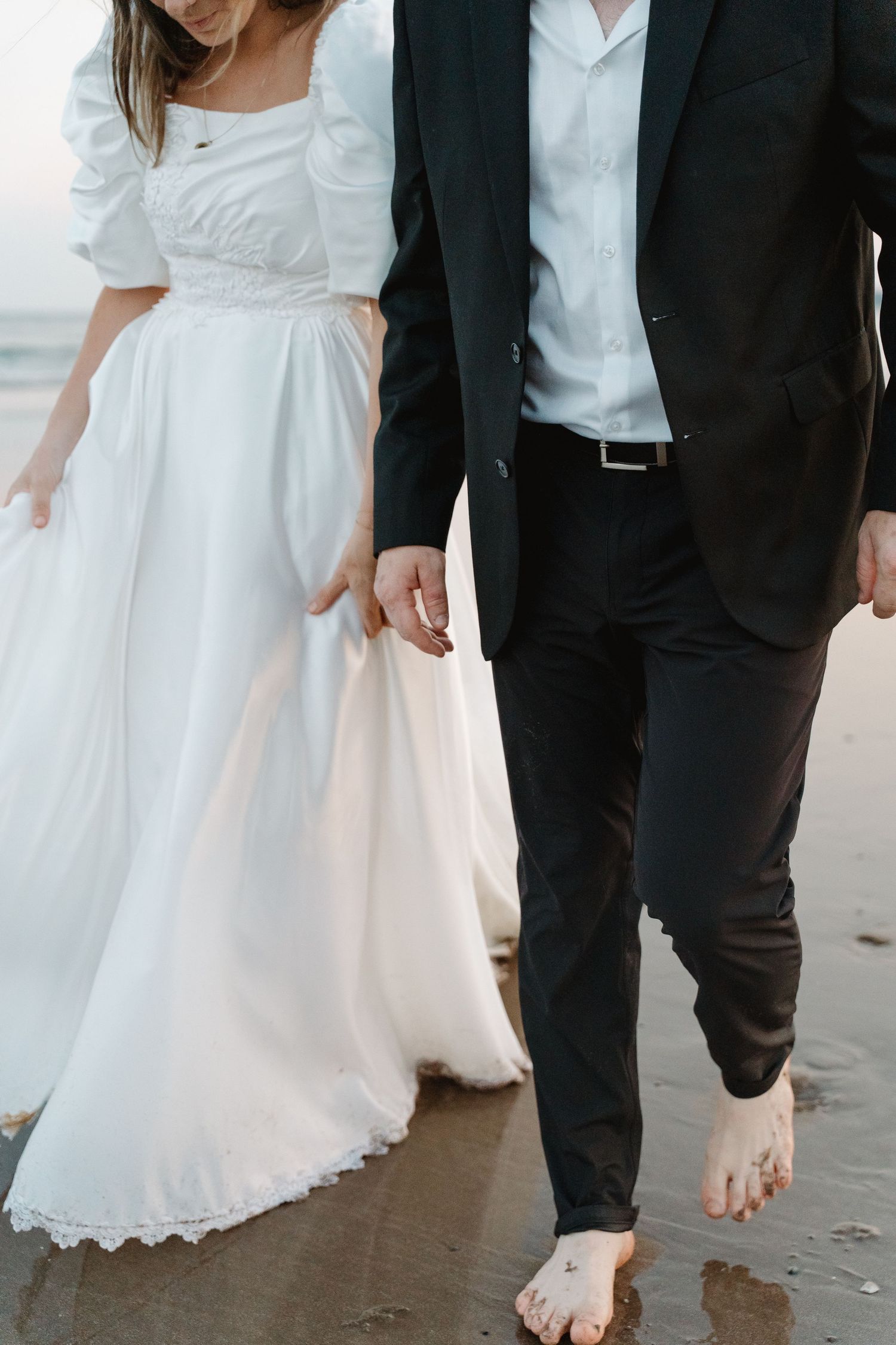 Bride in white lace dress and groom in black suit walking barefoot on sandy beach holding hands.