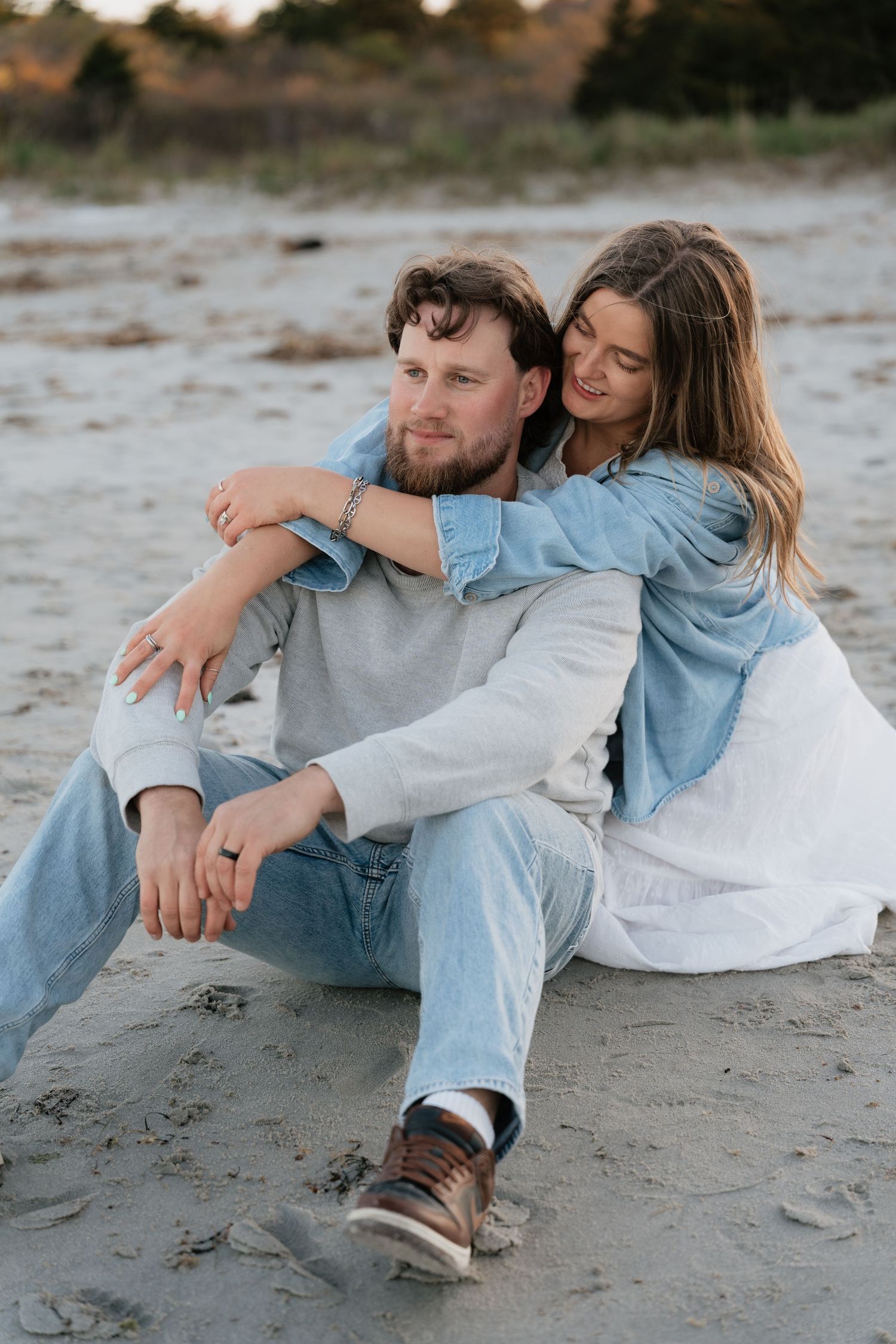 Couple sitting on sandy beach, woman embracing man from behind, both wearing casual denim outfits in soft natural light.