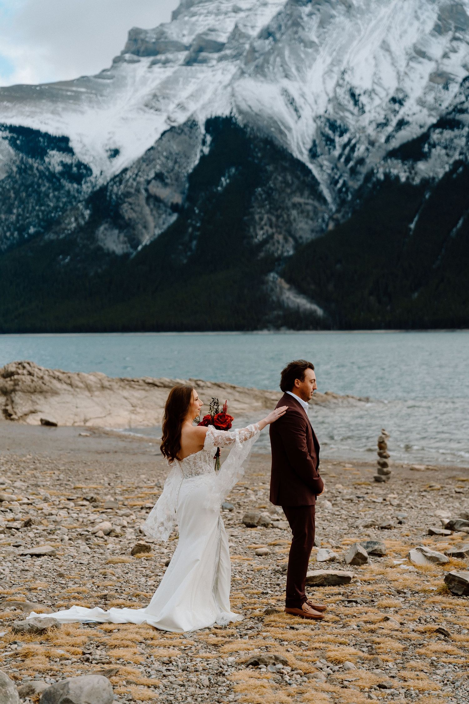 A couple in wedding attire dance together on a rocky shore with snow-capped mountains in the background.