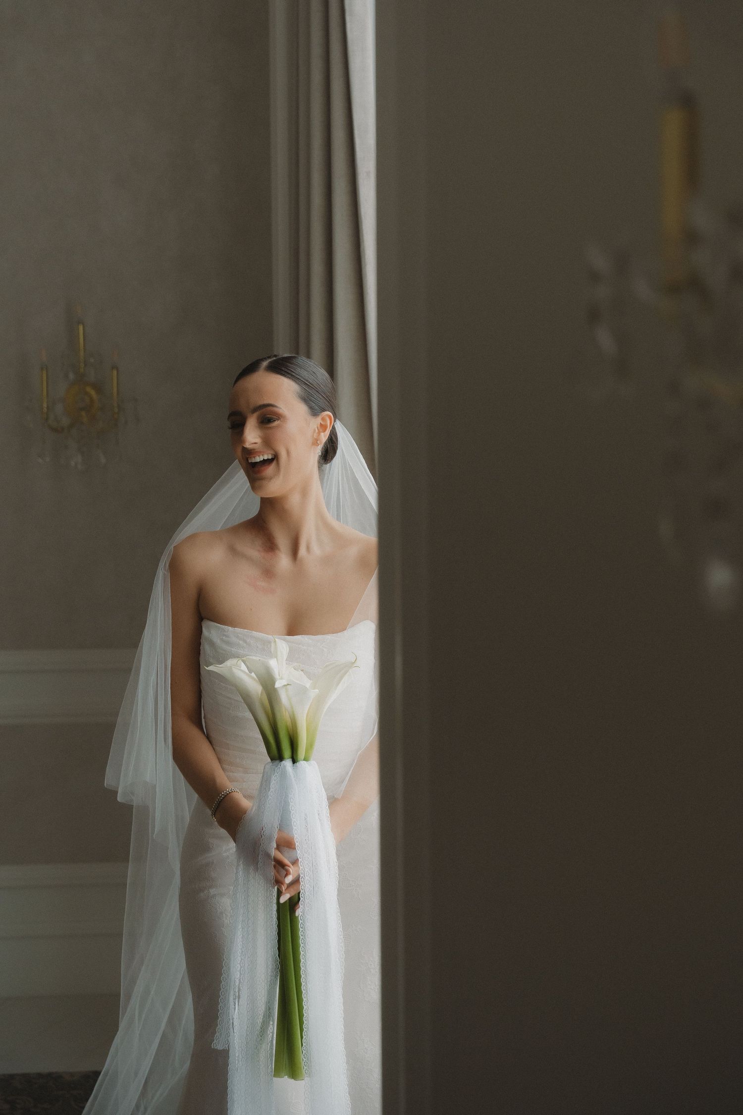 A bride in a classic white wedding dress and veil laughs joyfully during a candid moment before her ceremony.