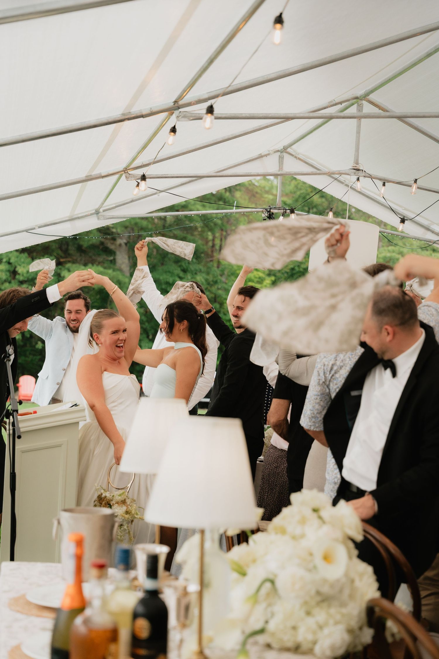 Bride celebrating joyfully with guests under a white tent with string lights at wedding reception.