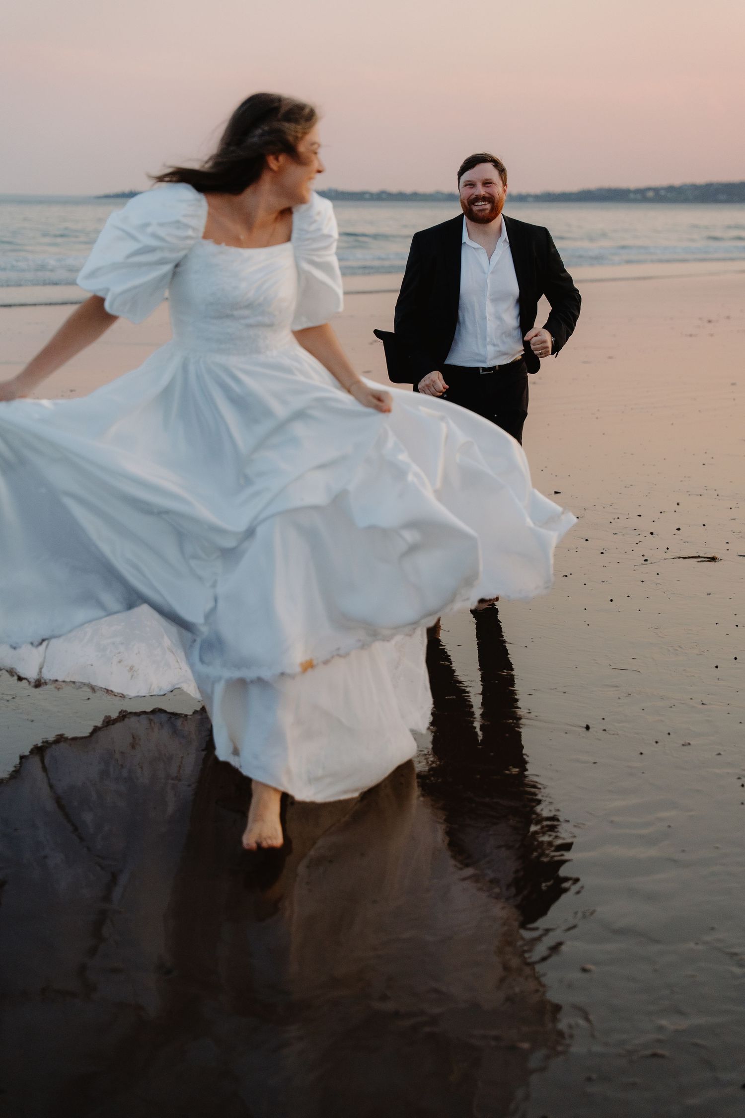 Bride in white dress runs on beach at sunset while groom in black suit smiles behind her near ocean waves.