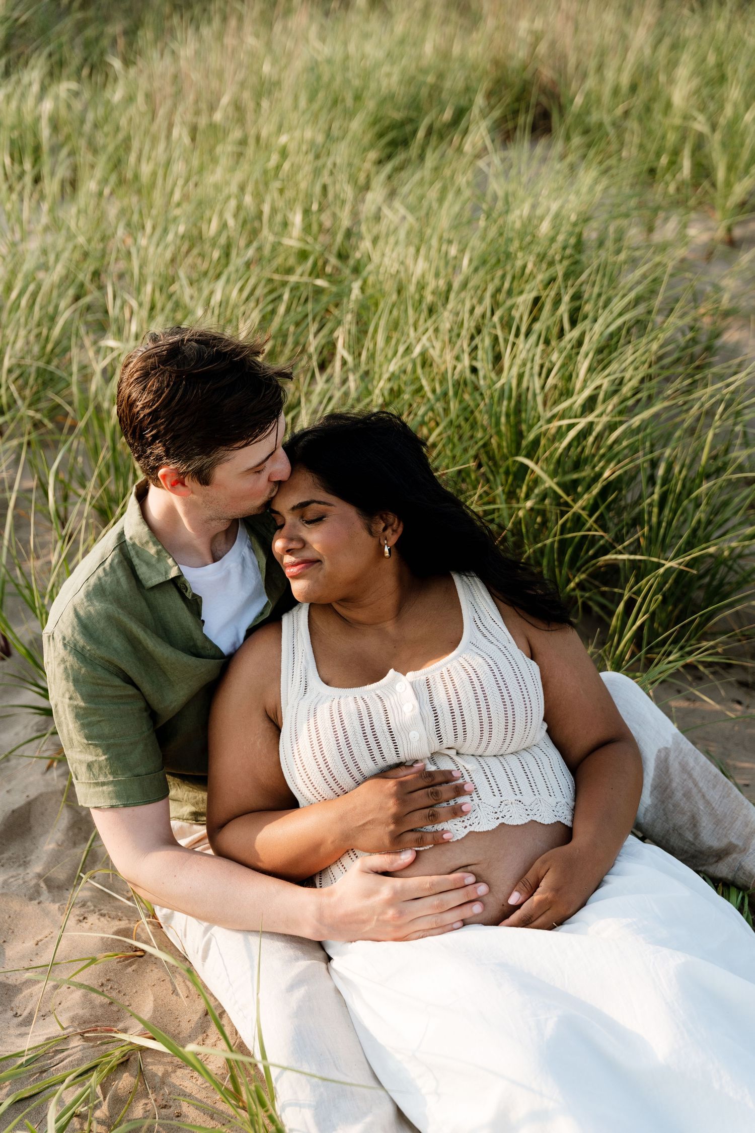 Couple sitting in beach grass, man kissing pregnant woman's temple while cradling her baby bump.