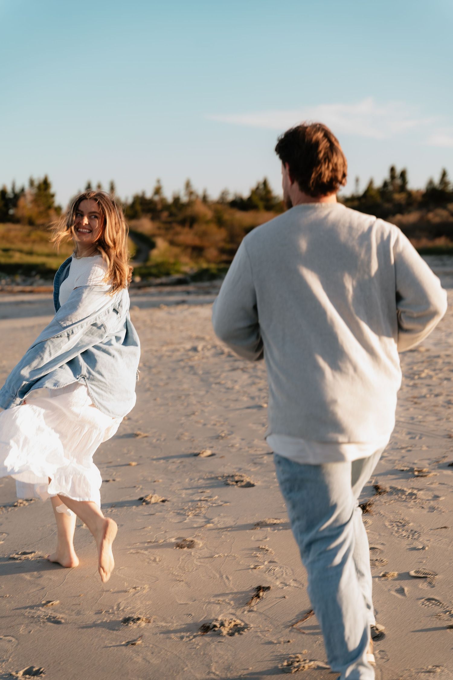 A couple playfully running on a sandy beach at golden hour, woman in white dress laughing.