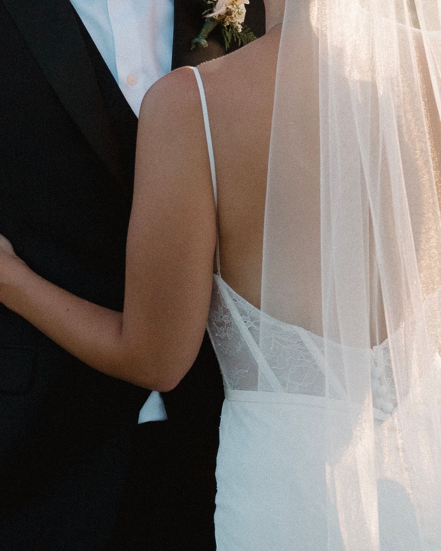 Close-up of bride in backless lace wedding gown with veil, embraced by groom in black tuxedo.