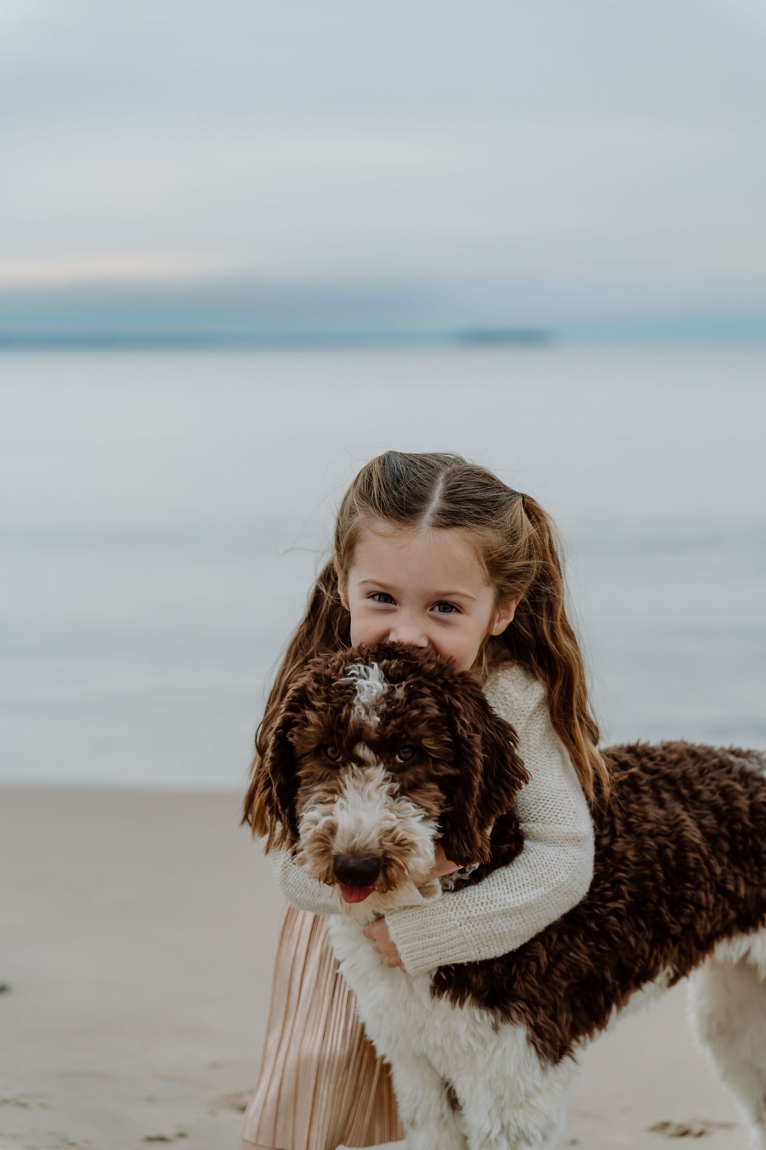 Child hugging brown and white dog on sandy beach with ocean backdrop.