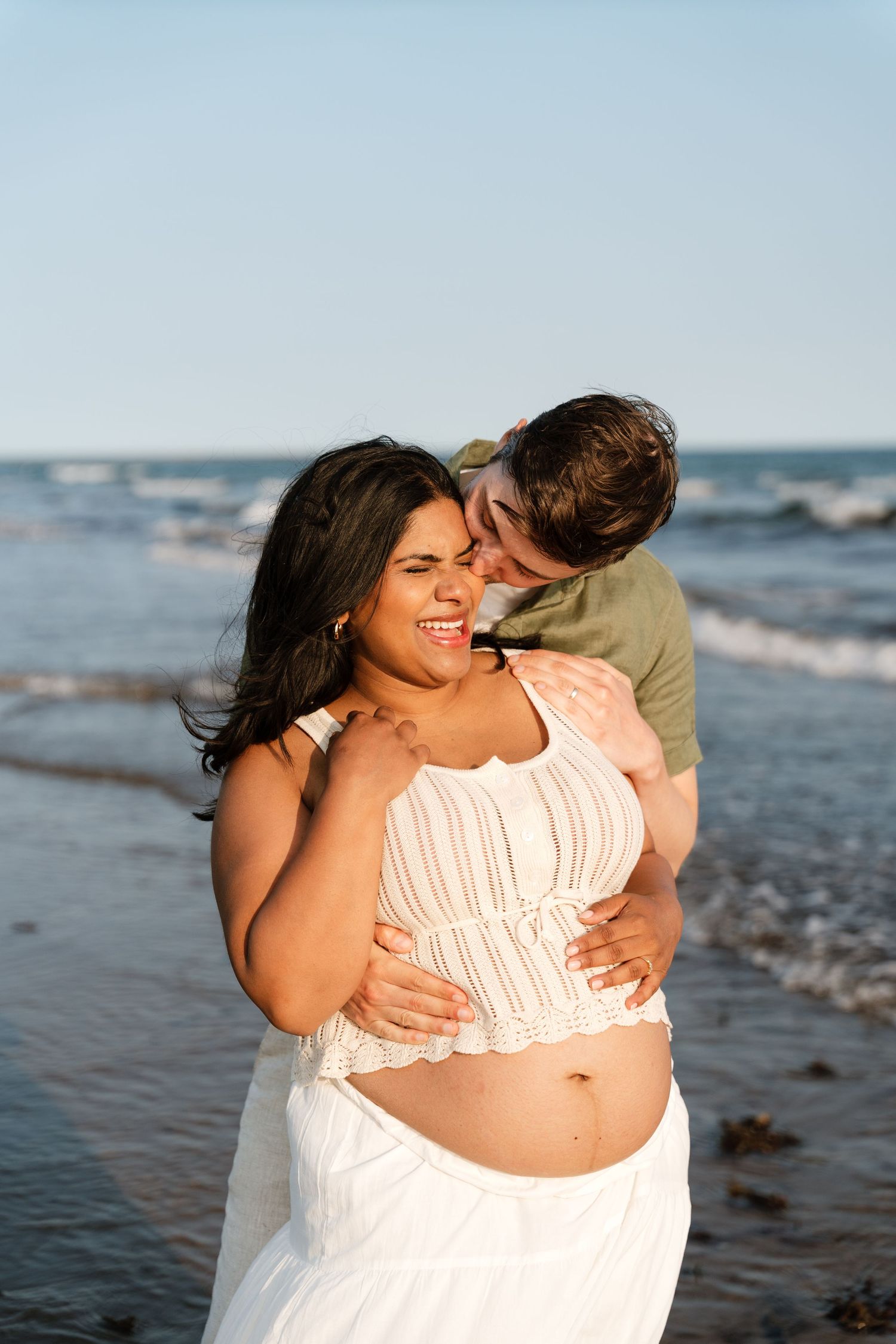 Pregnant couple embracing on beach at sunset, woman in white outfit laughing joyfully as man kisses her cheek.