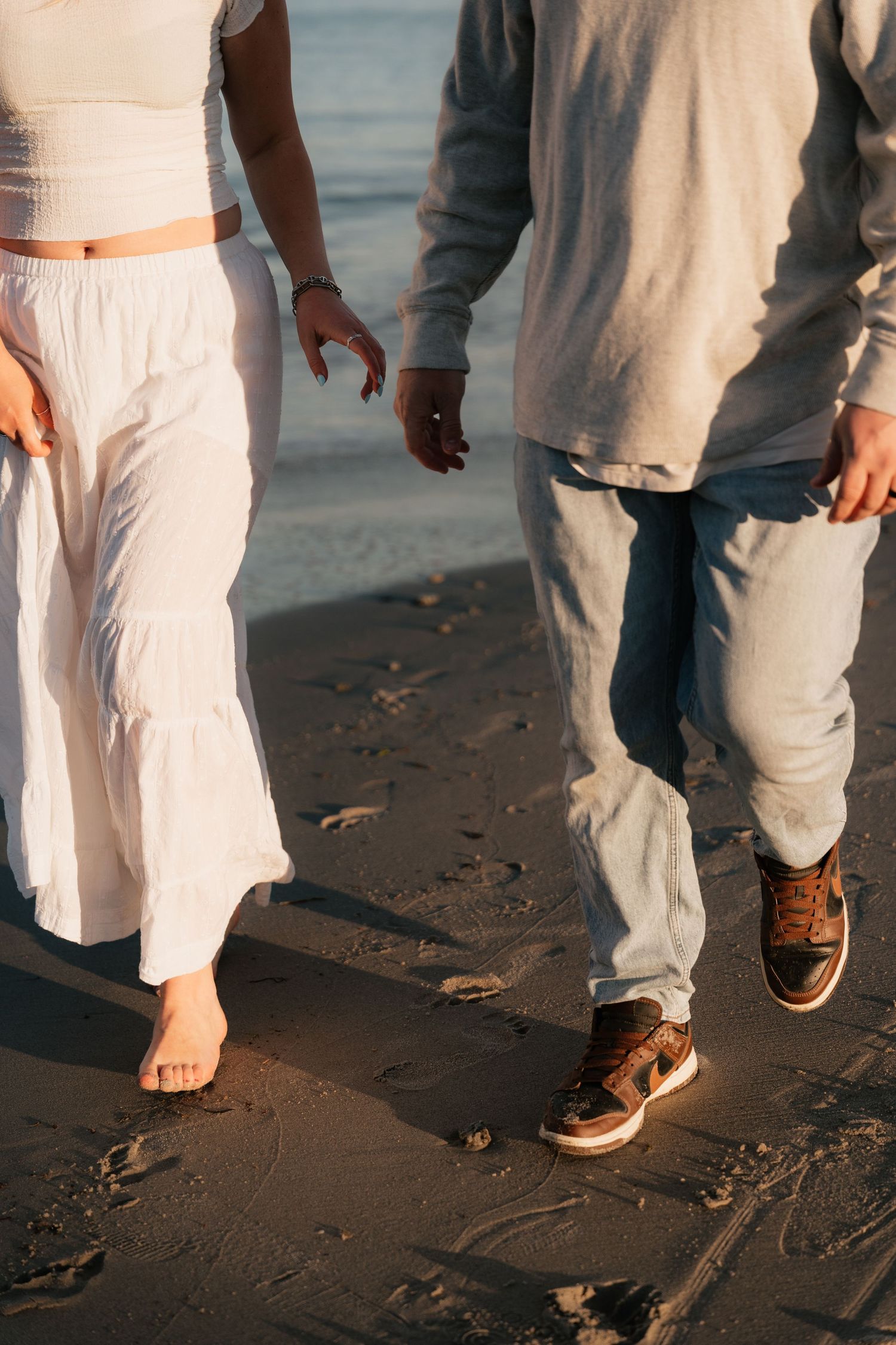 Couple walking barefoot on sandy beach at sunset, woman in white dress, man in jeans and sneakers.