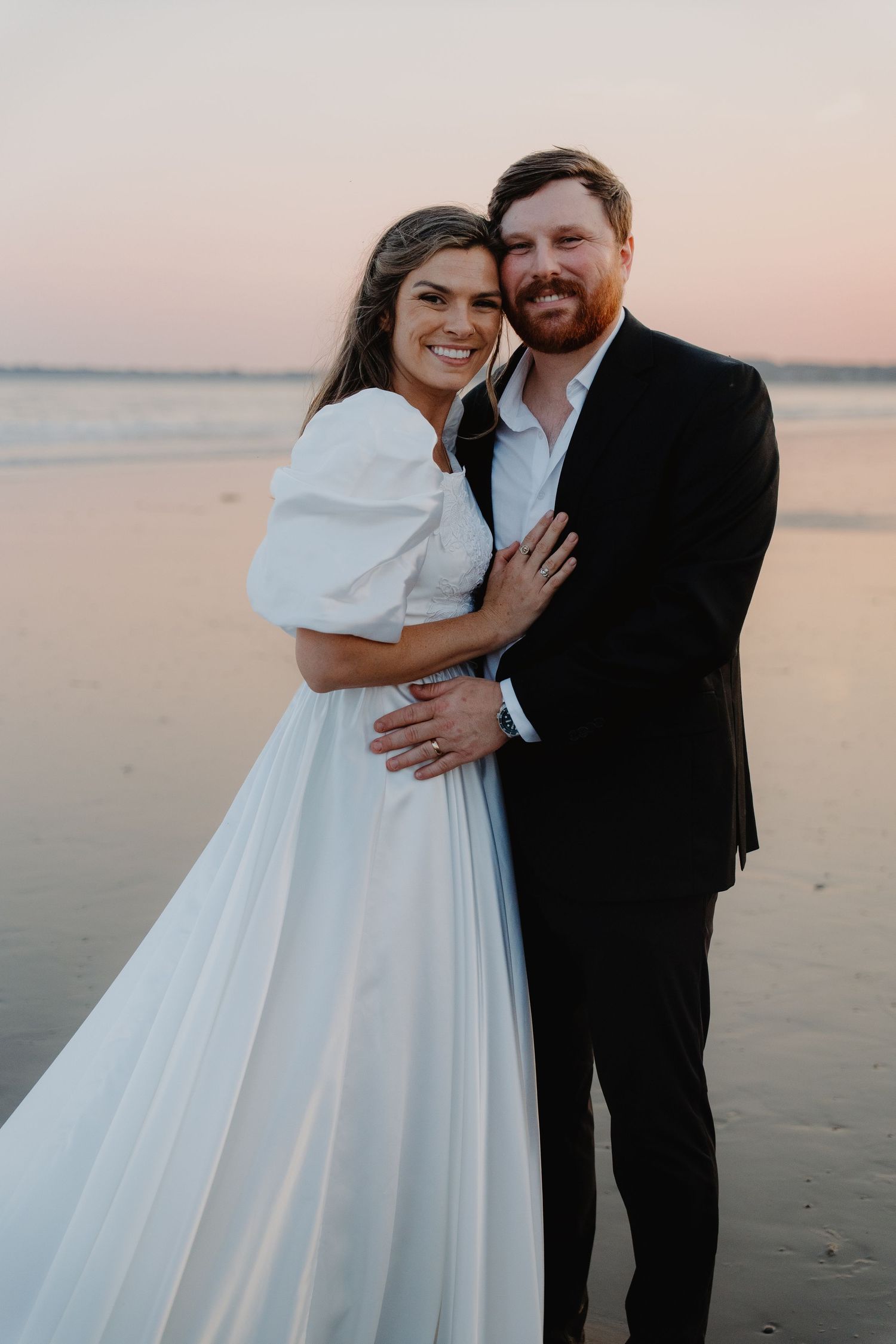 Bride in white puff-sleeve gown embraces groom in black suit on beach at sunset.