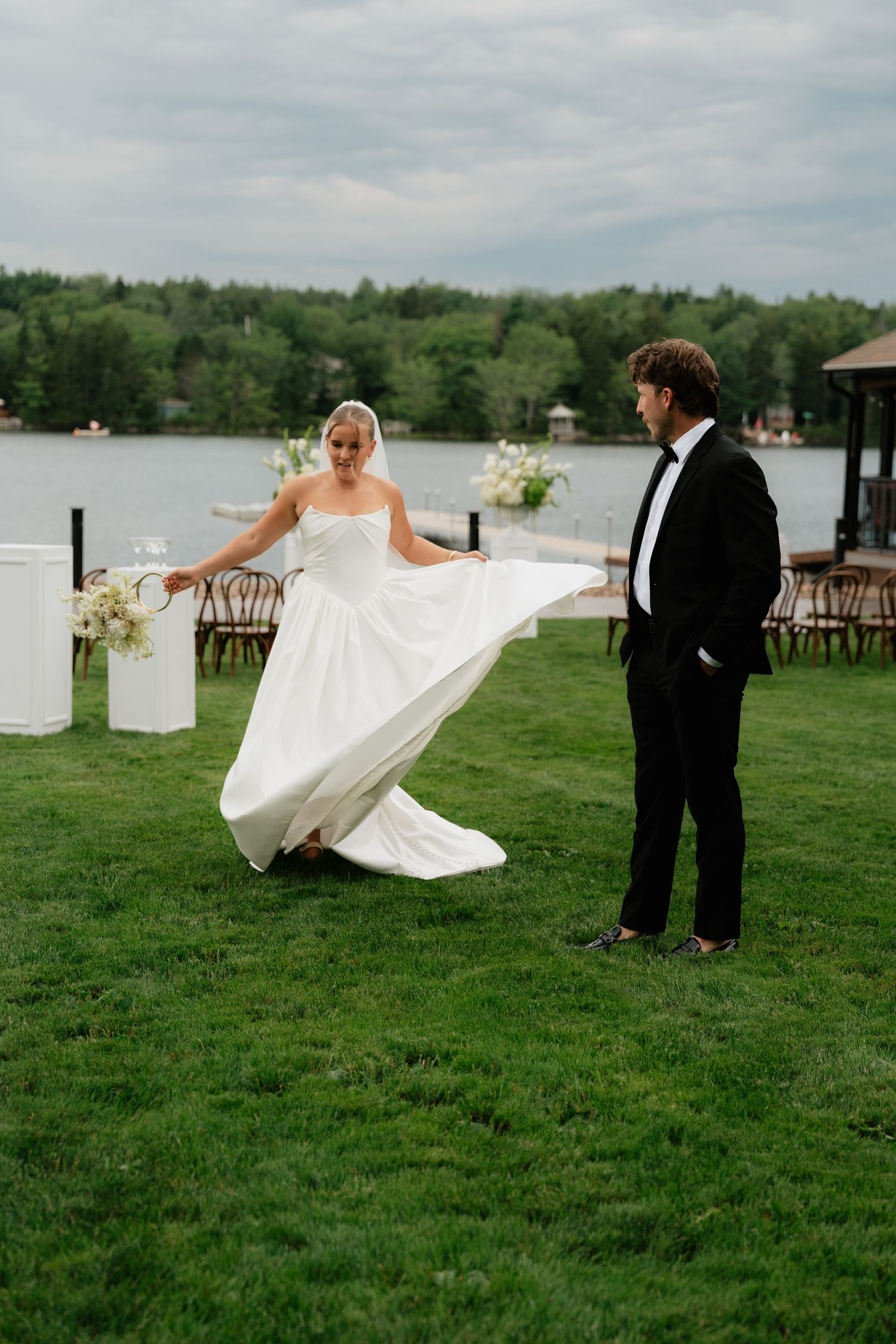 Bride twirling white gown on green lawn by lakeside wedding venue as groom in tuxedo watches.