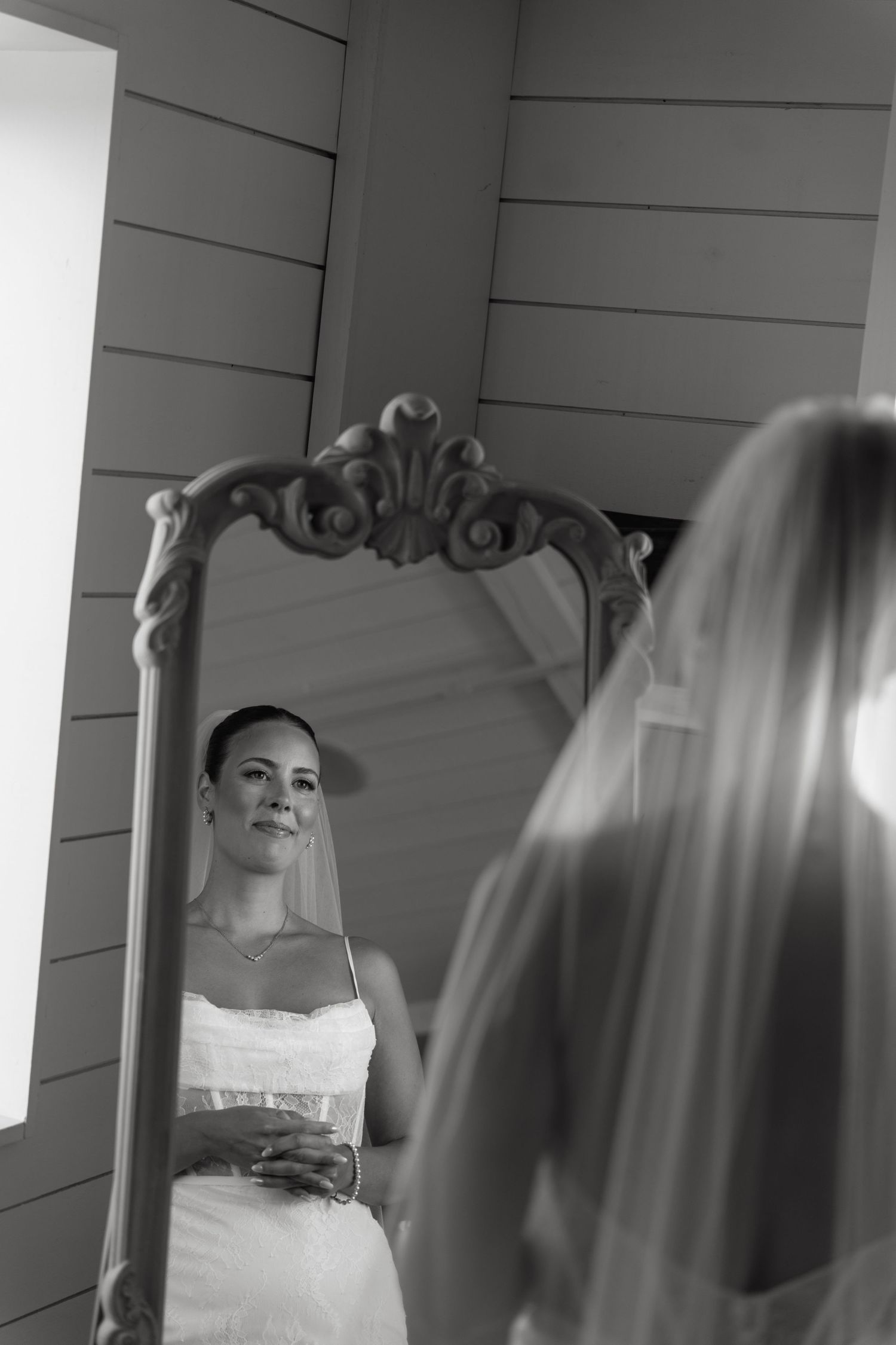 Black and white photo of a bride in a white dress and veil gazing at her reflection in an ornate vintage mirror.