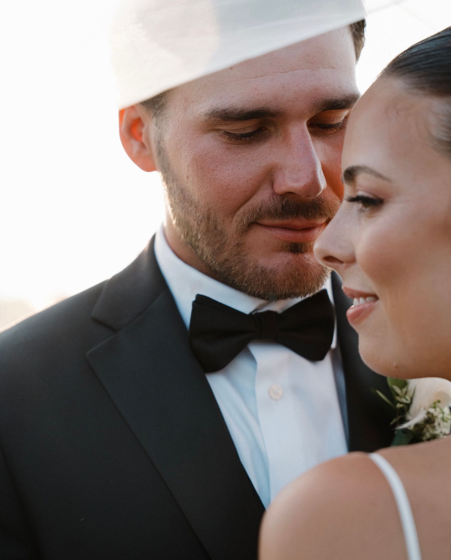 A groom in a black tuxedo and bow tie shares an intimate moment with his bride in a romantic wedding portrait.