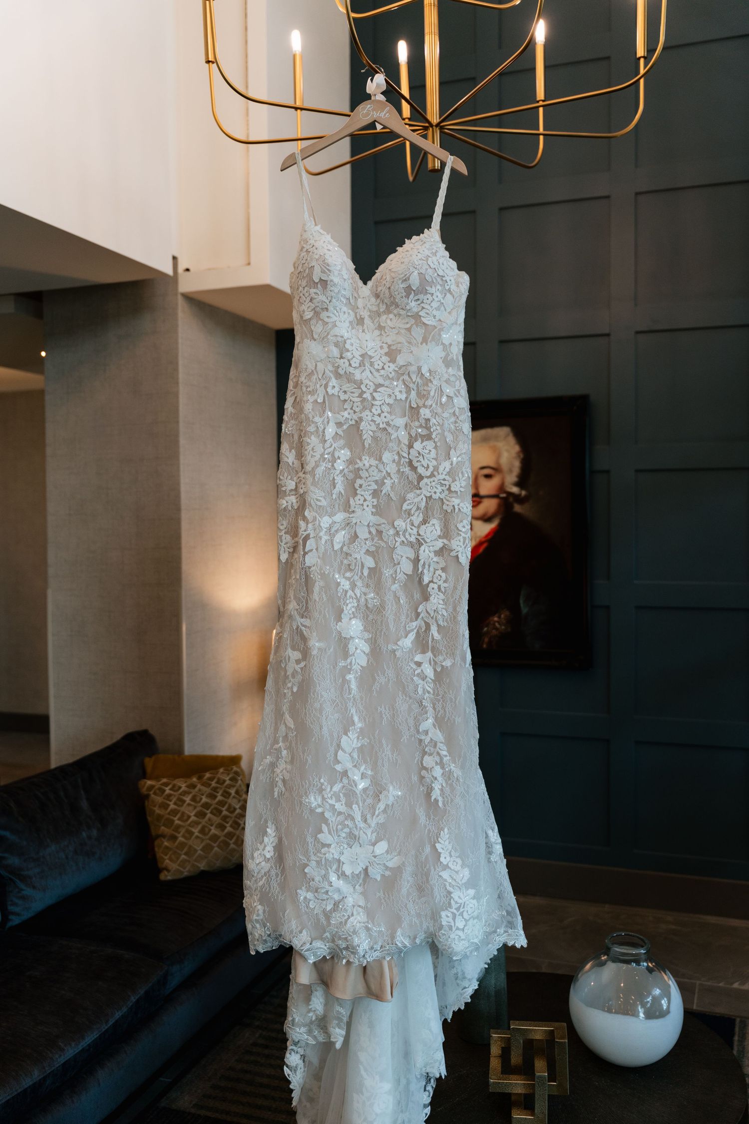 Elegant lace wedding dress hanging from a gold chandelier in a stylish hotel suite with dark green paneled walls.