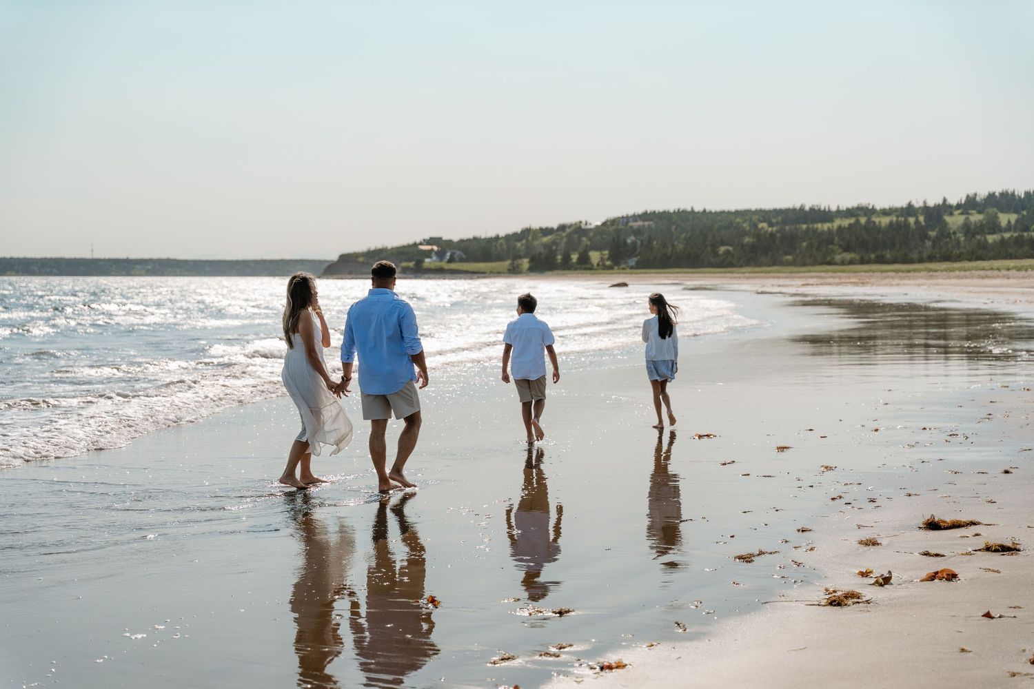 Family of four walking along a sandy beach shoreline with gentle waves and a tree-lined coast in the background.