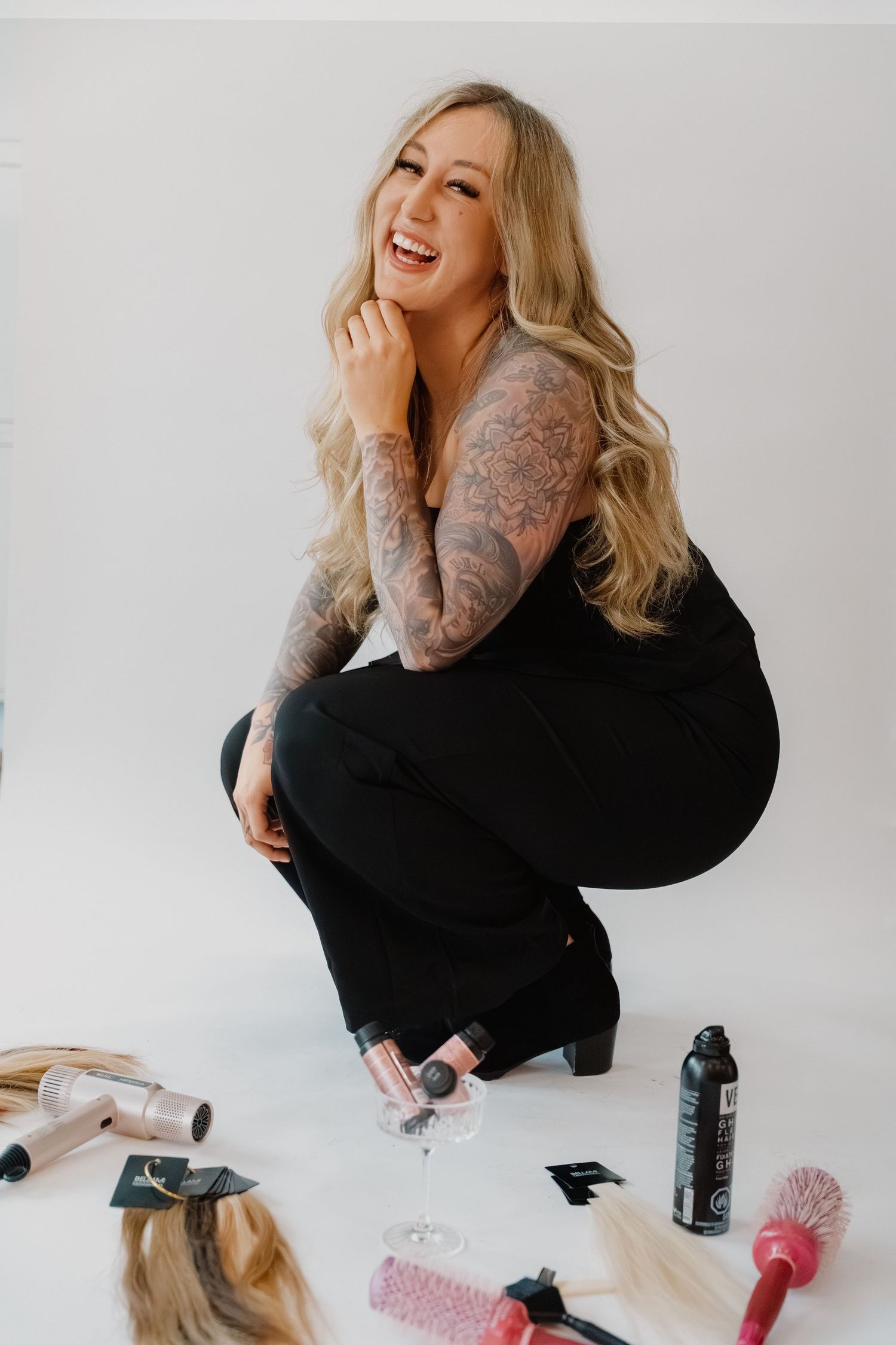 Smiling blonde woman with sleeve tattoos crouching in black outfit surrounded by hair tools and products.