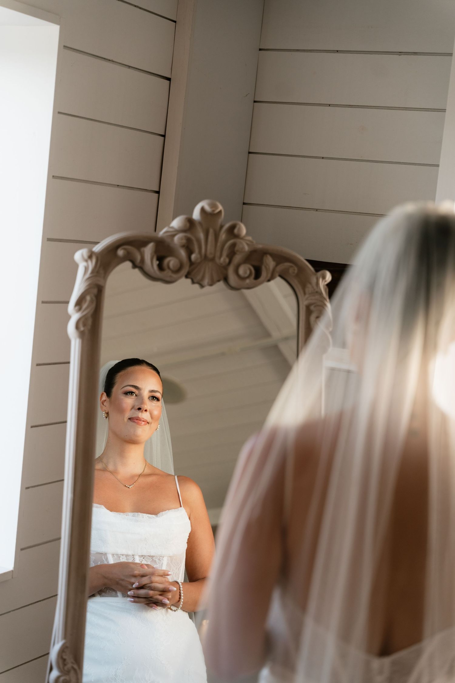 Bride in white gown and veil gazes at her reflection in an ornate vintage mirror while getting ready.