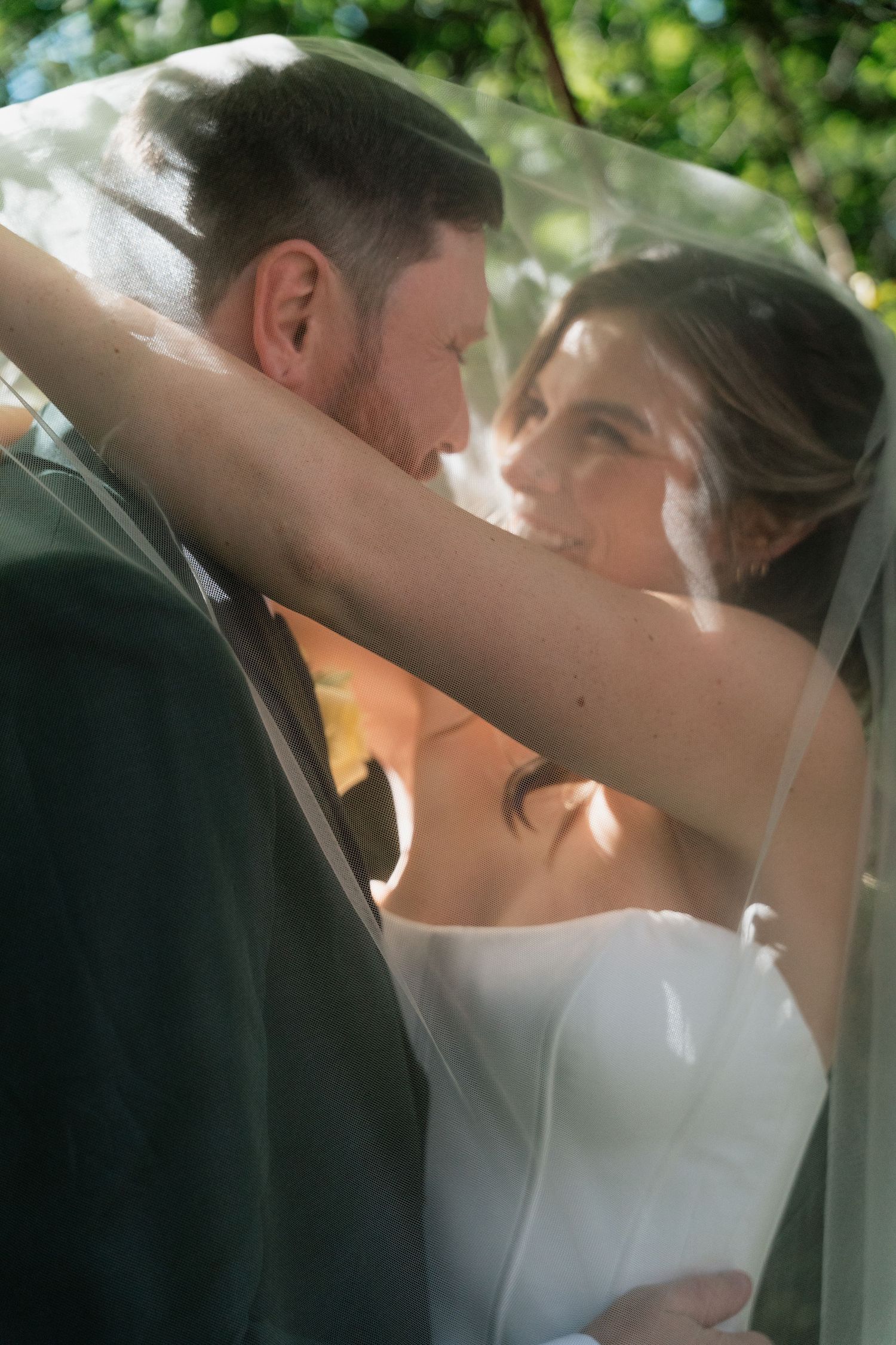 Bride and groom share an intimate moment under a sheer wedding veil, surrounded by lush green trees.